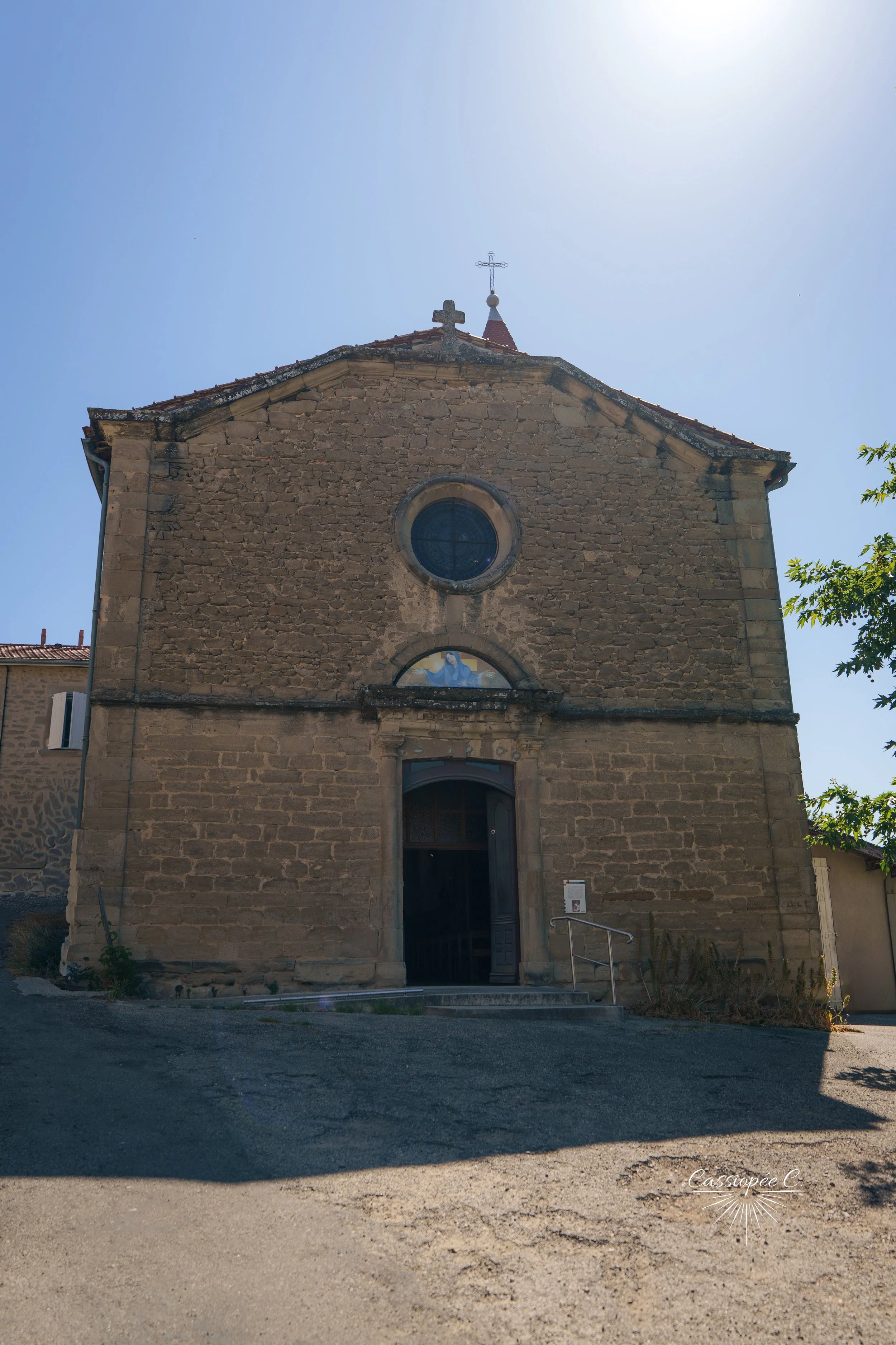 Façade en pierre d'une ancienne église avec un rosace, une porte en bois et un petit panneau d'information devant. Le ciel est clair et ensoleillé.