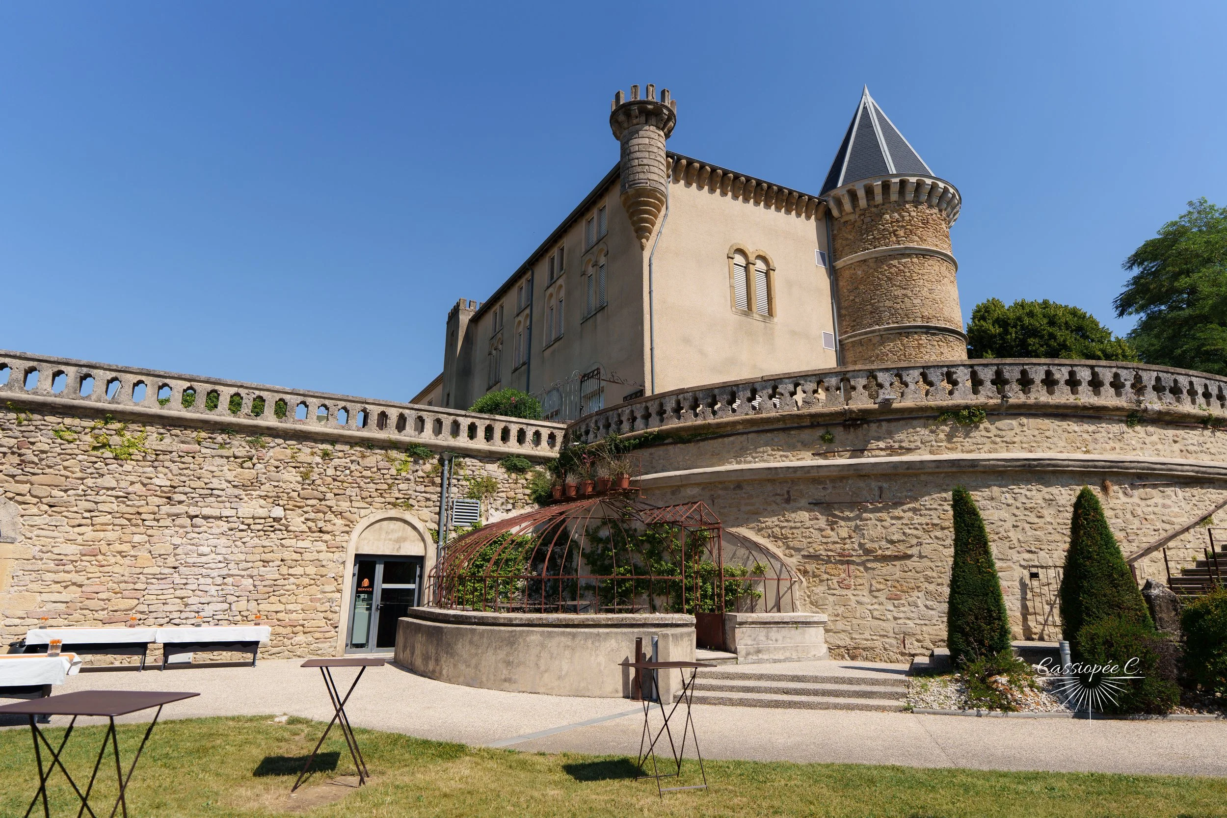 Un château avec une tour ronde en pierre et une tour conique, entouré d'une terrasse en pierre avec une balustrade. Au premier plan, un jardin avec des petits arbres et une zone de terrasse avec des tables, sous un ciel bleu dégagé.