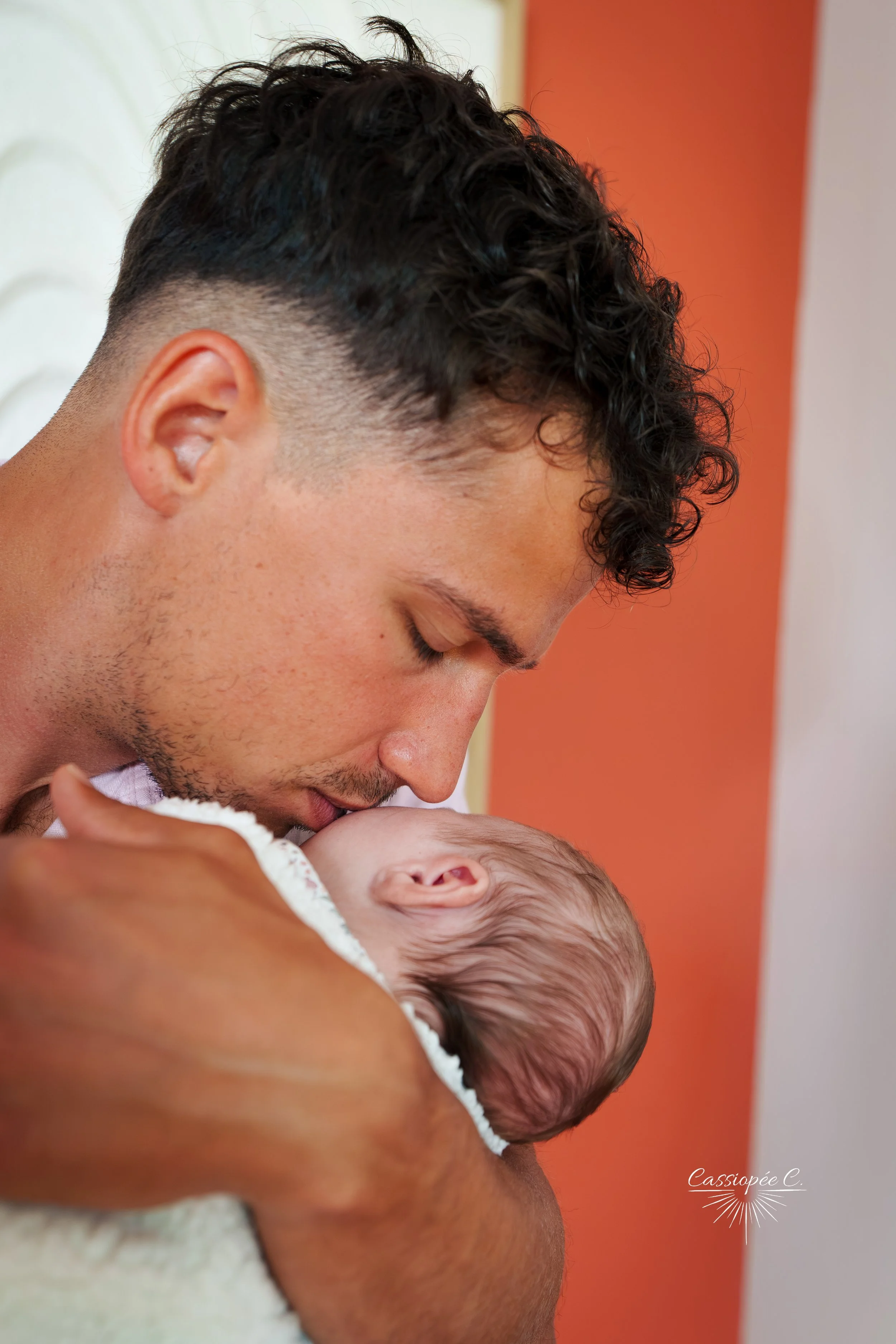 Un homme jeune avec des cheveux bouclés noir fait un câlin à un bébé, dans un environnement intérieur avec des murs orange et blanc