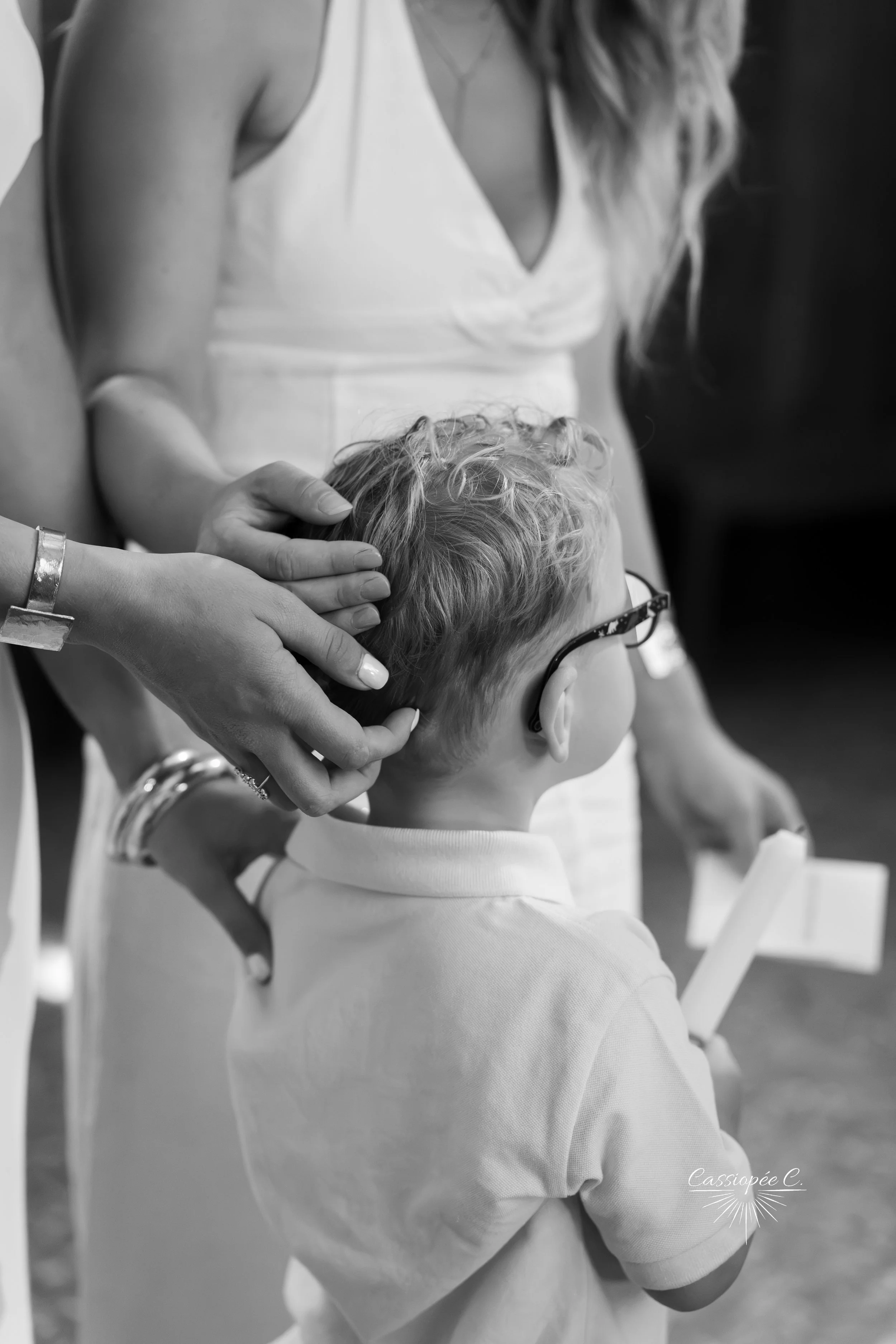 Enfant avec lunettes assis, femme touchant ses cheveux, en intérieur, en noir et blanc.