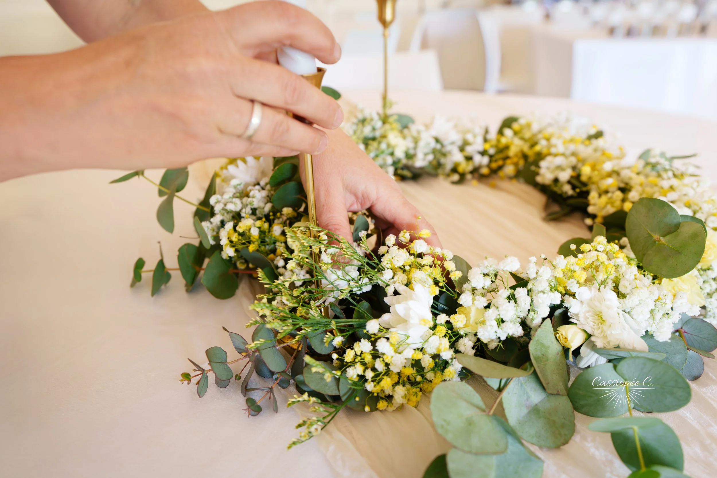 Une main ajuste un feuillage dans une composition florale composée de petites fleurs blanches et jaunes et de feuilles vertes, posée sur une table en bois.