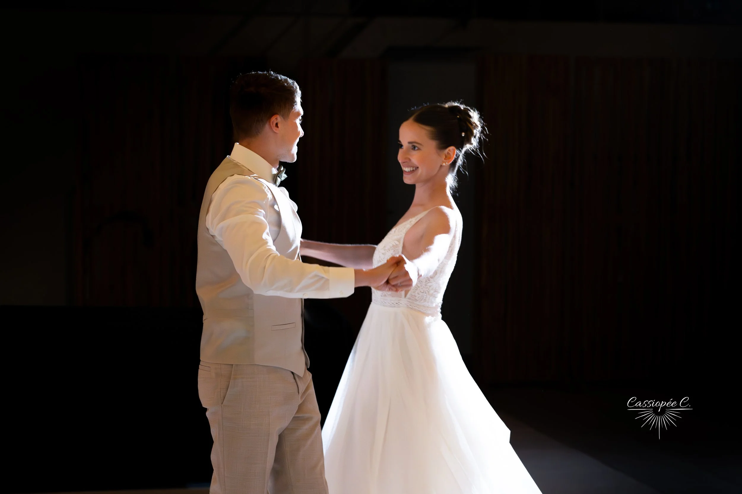 Un couple danse lors d'un mariage, la femme porte une robe de mariée blanche et l'homme un costume clair, dans une salle sombre avec un éclairage focalisé.