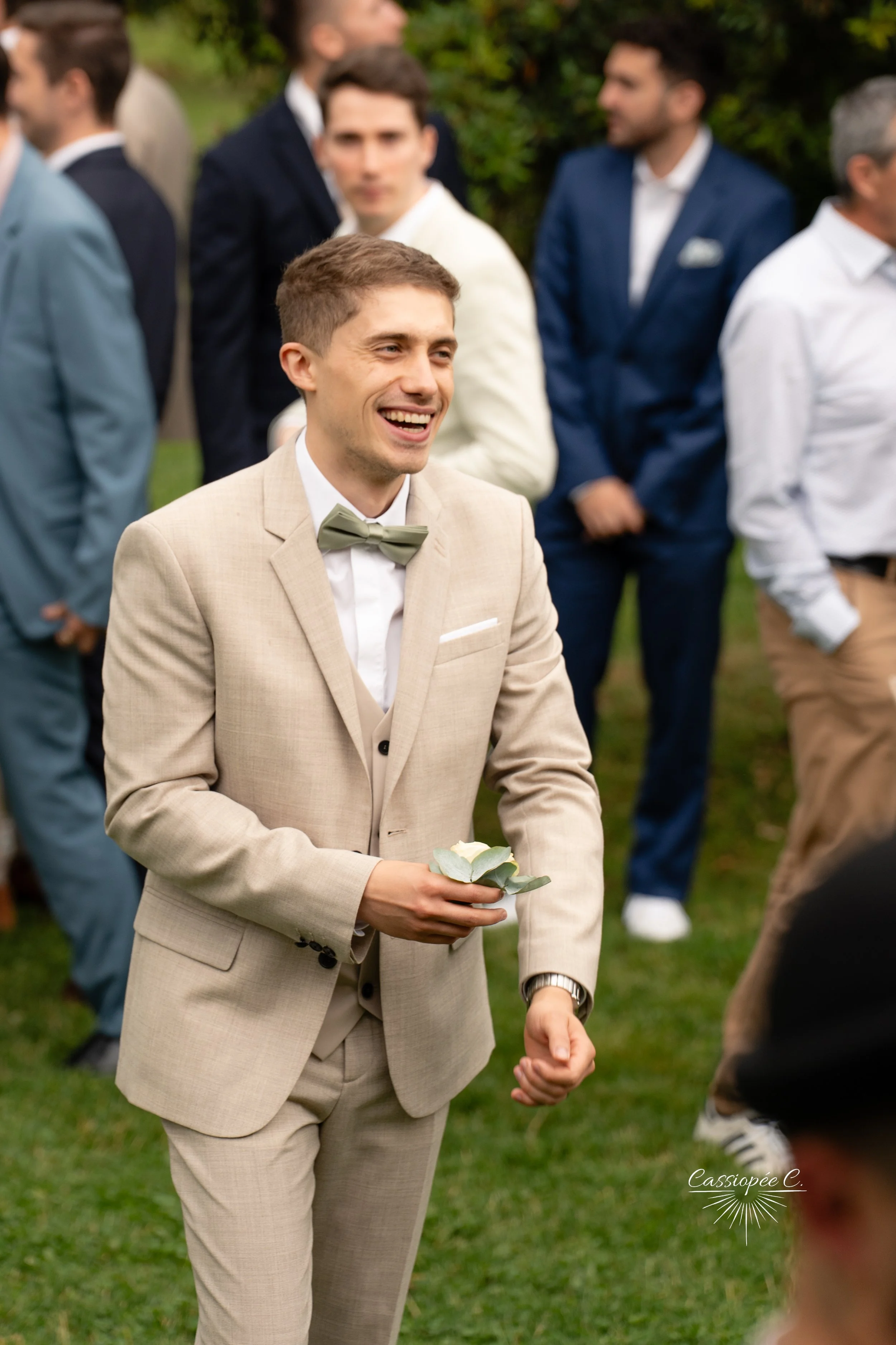 Le marié souriant vêtu d'un costume beige avec un nœud papillon vert, tenant une boutonnière, lors d'une cérémonie en plein air, entouré d'autres hommes en costume.