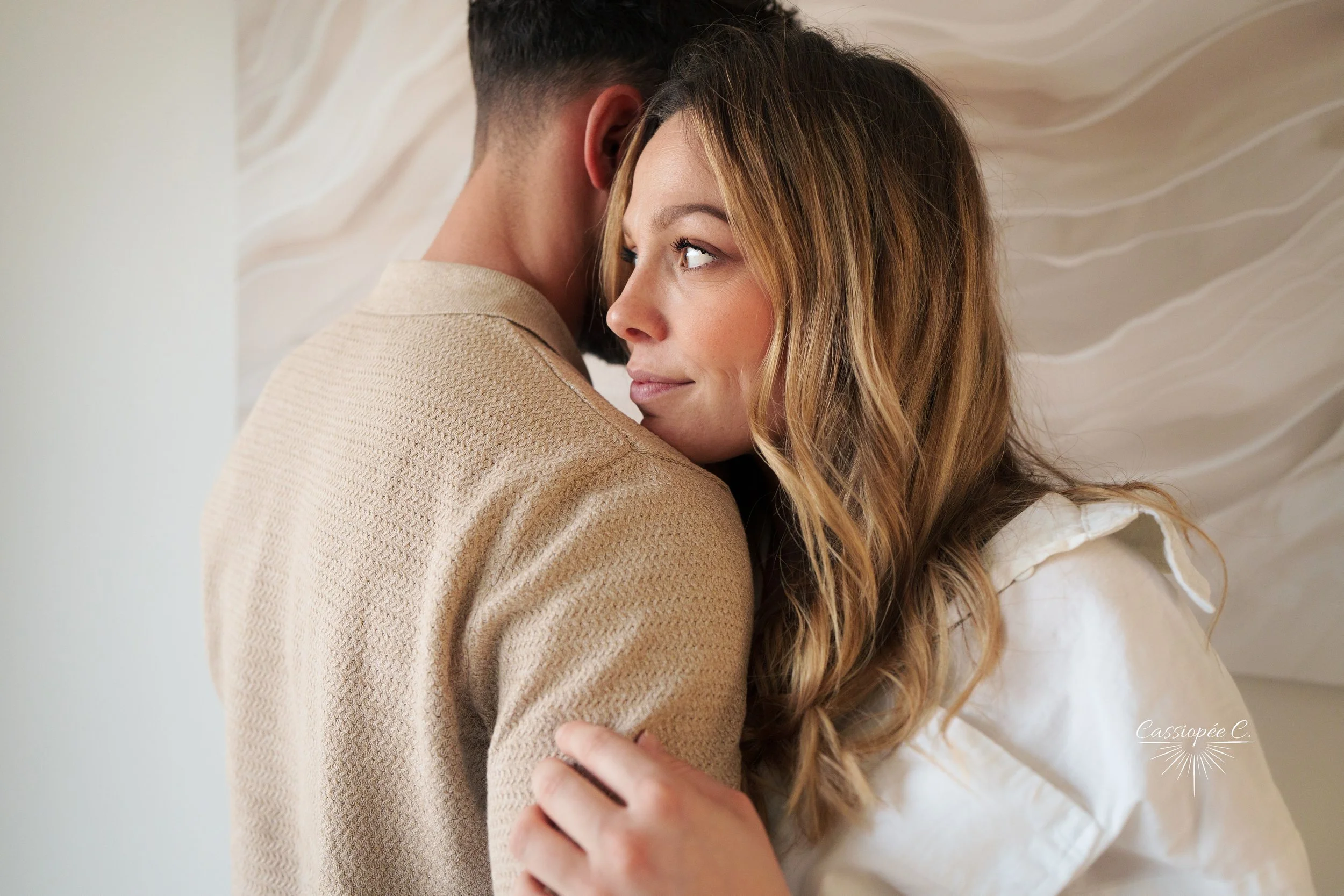 Un couple en étreinte, la femme regarde le côté droit. L’arrière-plan présente un mur blanc avec un motif texturé.