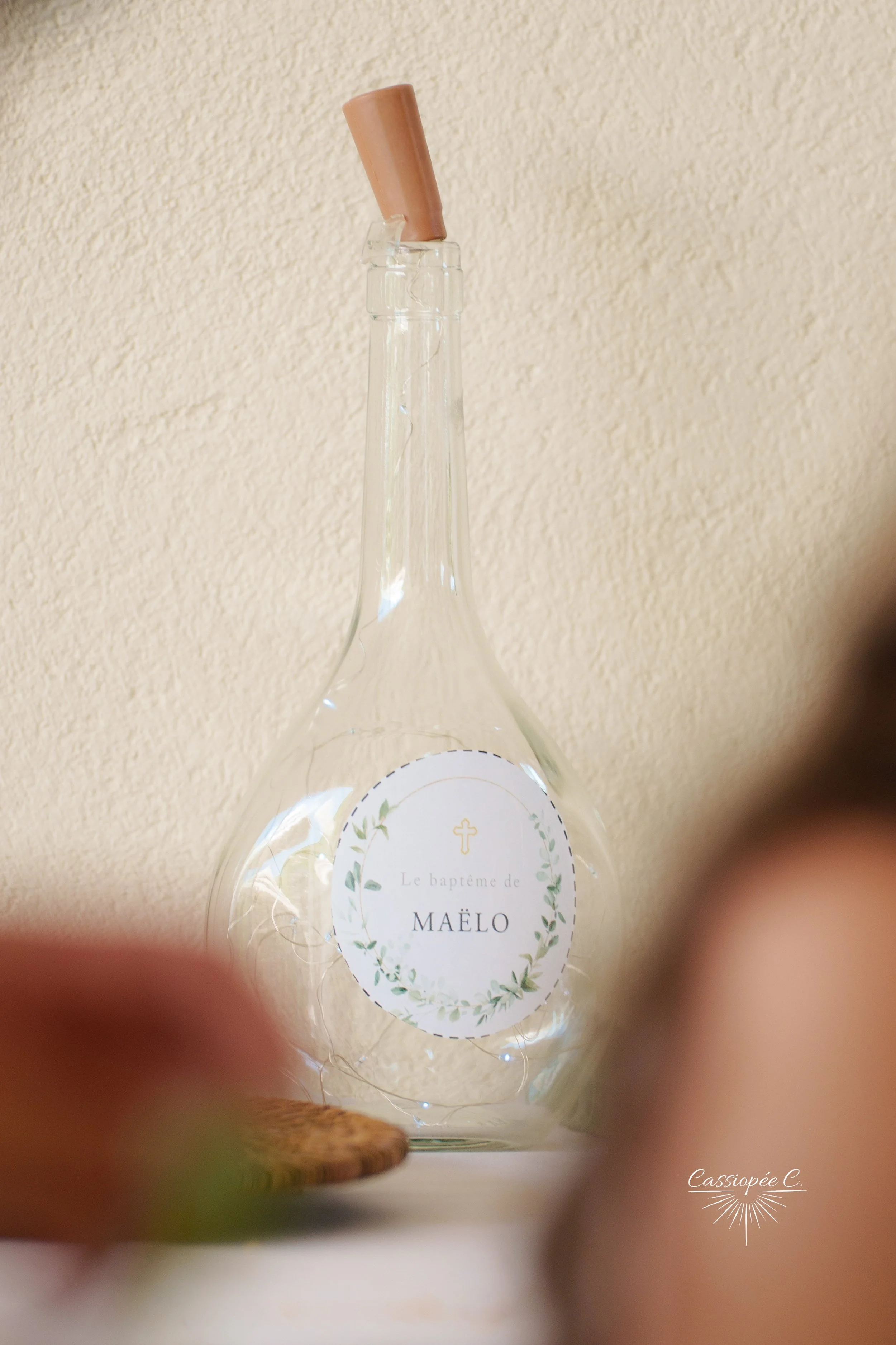 Flacon en verre transparent avec une étiquette indiquant le baptême de Maëlo, décorée de feuilles vertes et d'une petite croix.