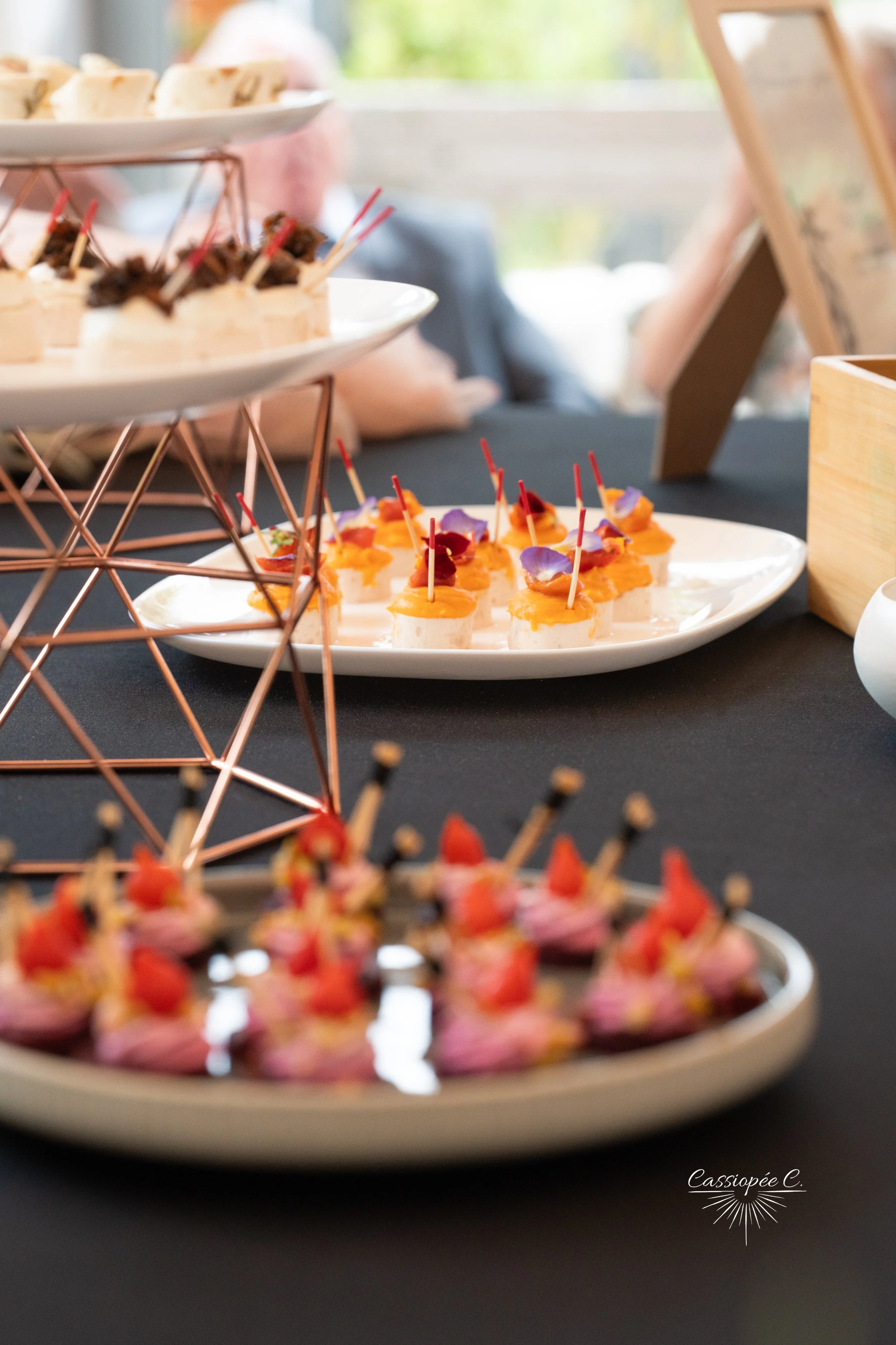 Plateaux de petits fours et amuse-bouches décorés avec des fleurs et des pique en bois, présentation élégante sur une table noire dans un environnement lumineux.