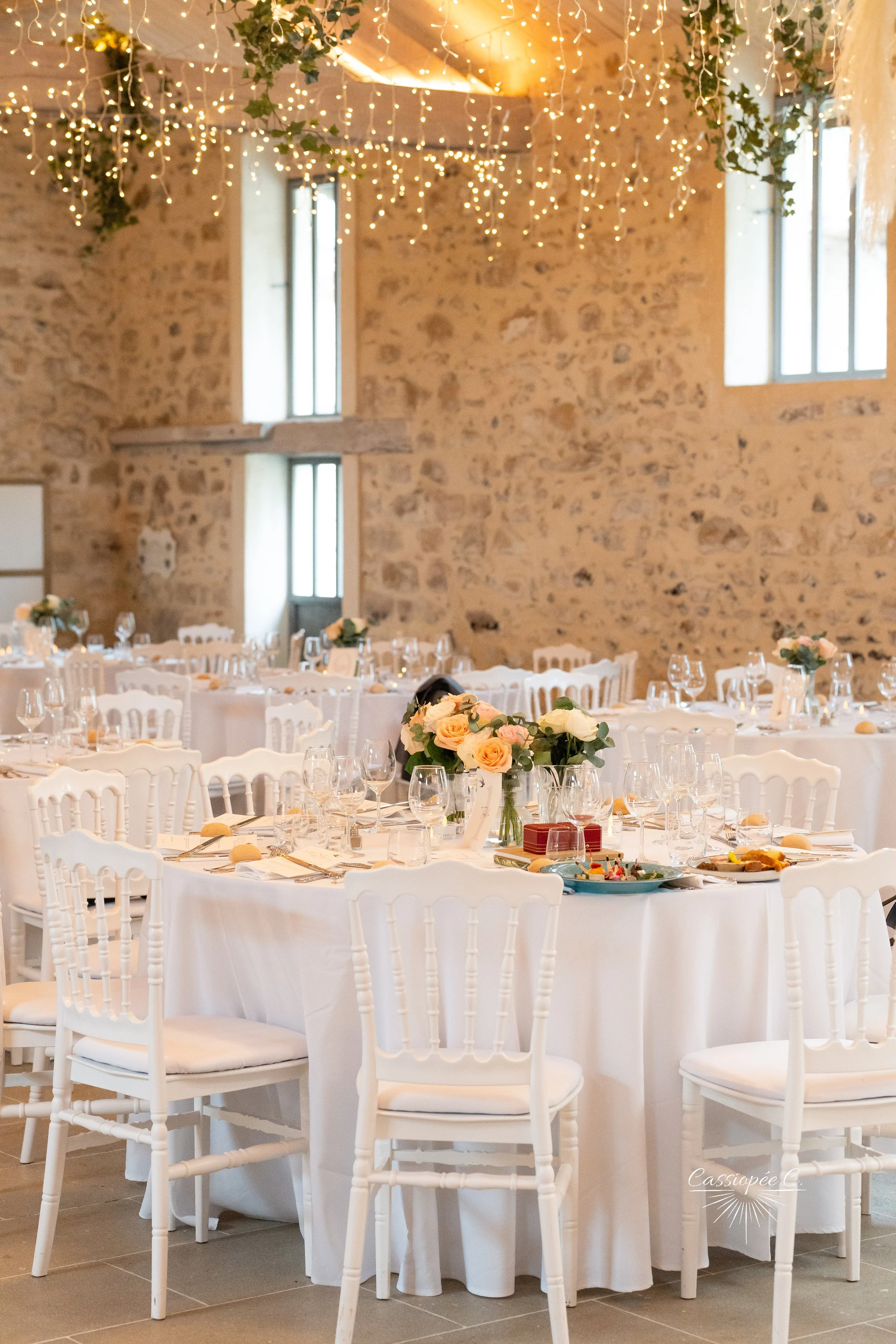 Salle de mariage décorée avec des tables rondes drapées de blanc, des chaises blanches, des arrangements de fleurs, et des guirlandes lumineuses suspendues au plafond. Le mur de fond est en pierre.