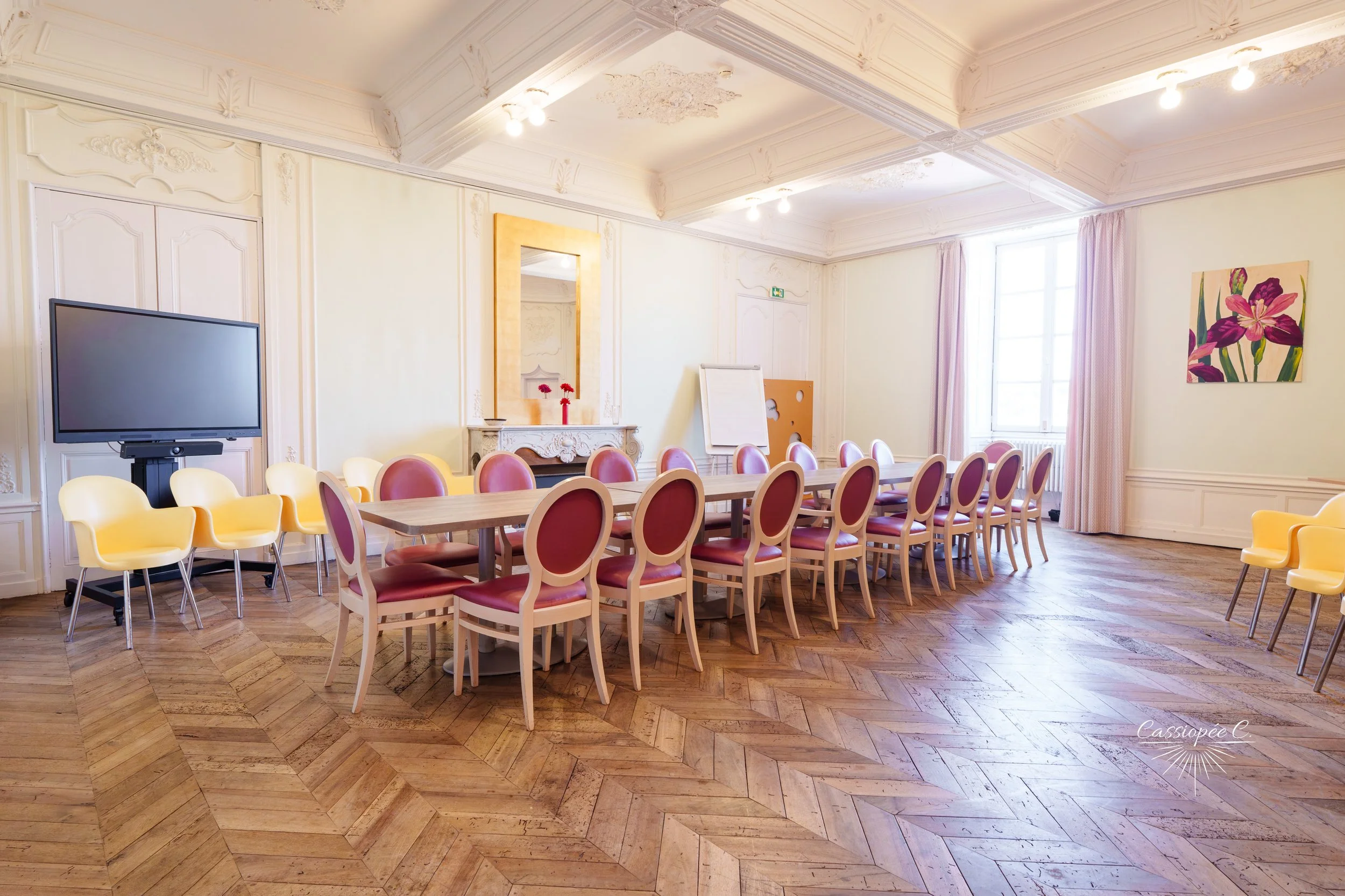 Salle de réunion avec une grande table entourée de chaises roses et jaunes, un grand écran à gauche, un grand miroir doré au fond, une peinture de fleurs roses et mauves à droite, fenêtres avec rideaux roses, plafond orné avec moulures et éclairage