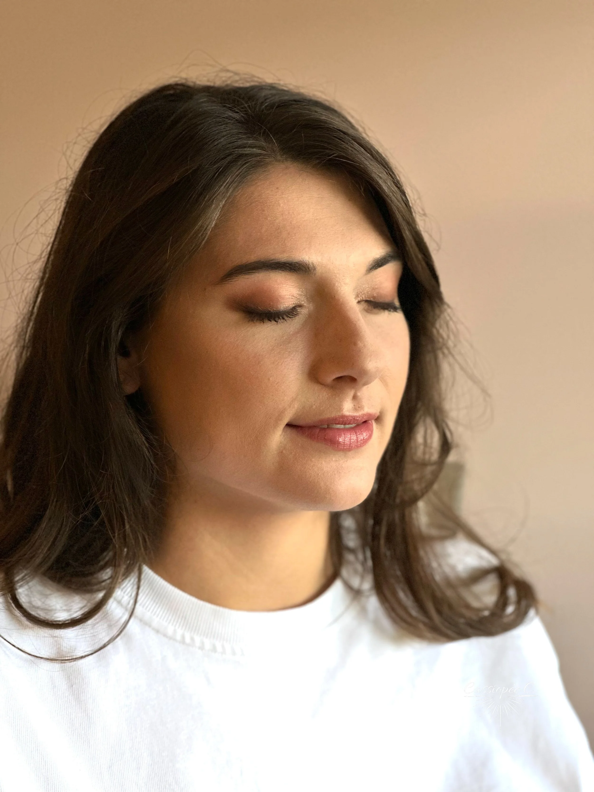 Un portrait d'une femme aux yeux fermés, avec des cheveux longs, bruns et ondulés, portant un t-shirt blanc, maquillage pour un mariage.