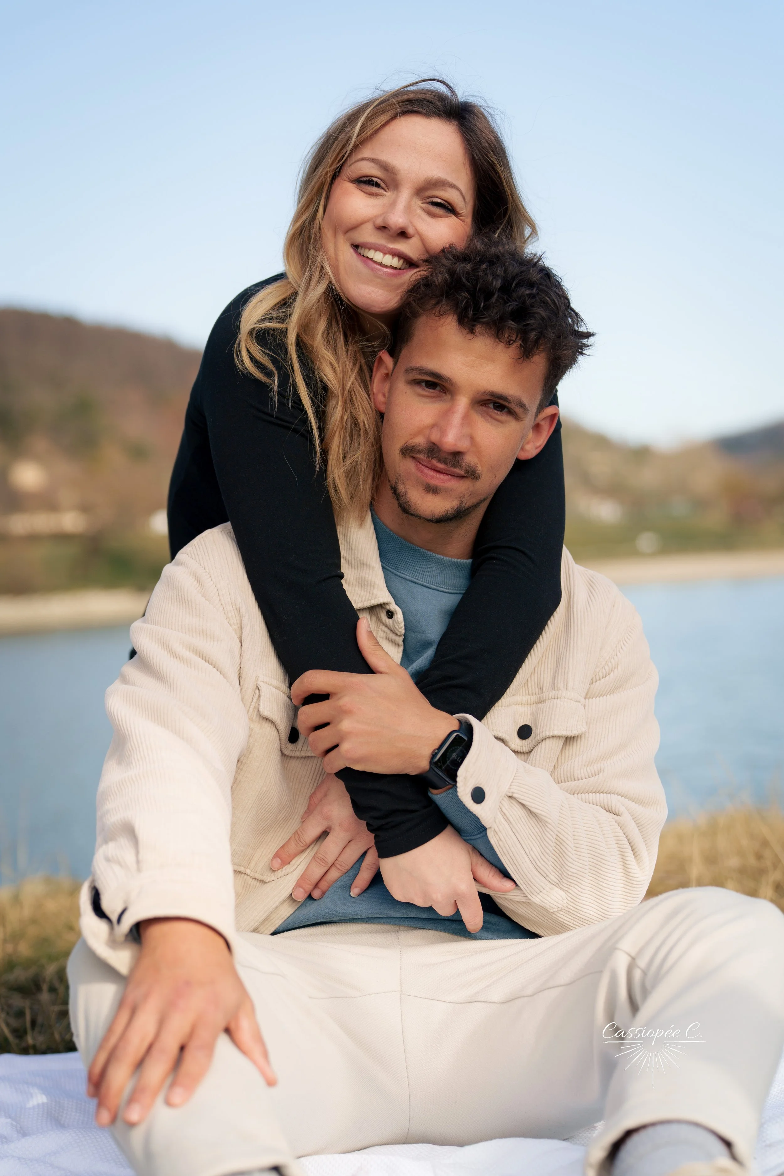 Un couple souriant assis près d'un lac avec un arrière-plan de collines et un ciel clair, l'homme portant une veste beige et la femme portant un pull noir, en extérieur.