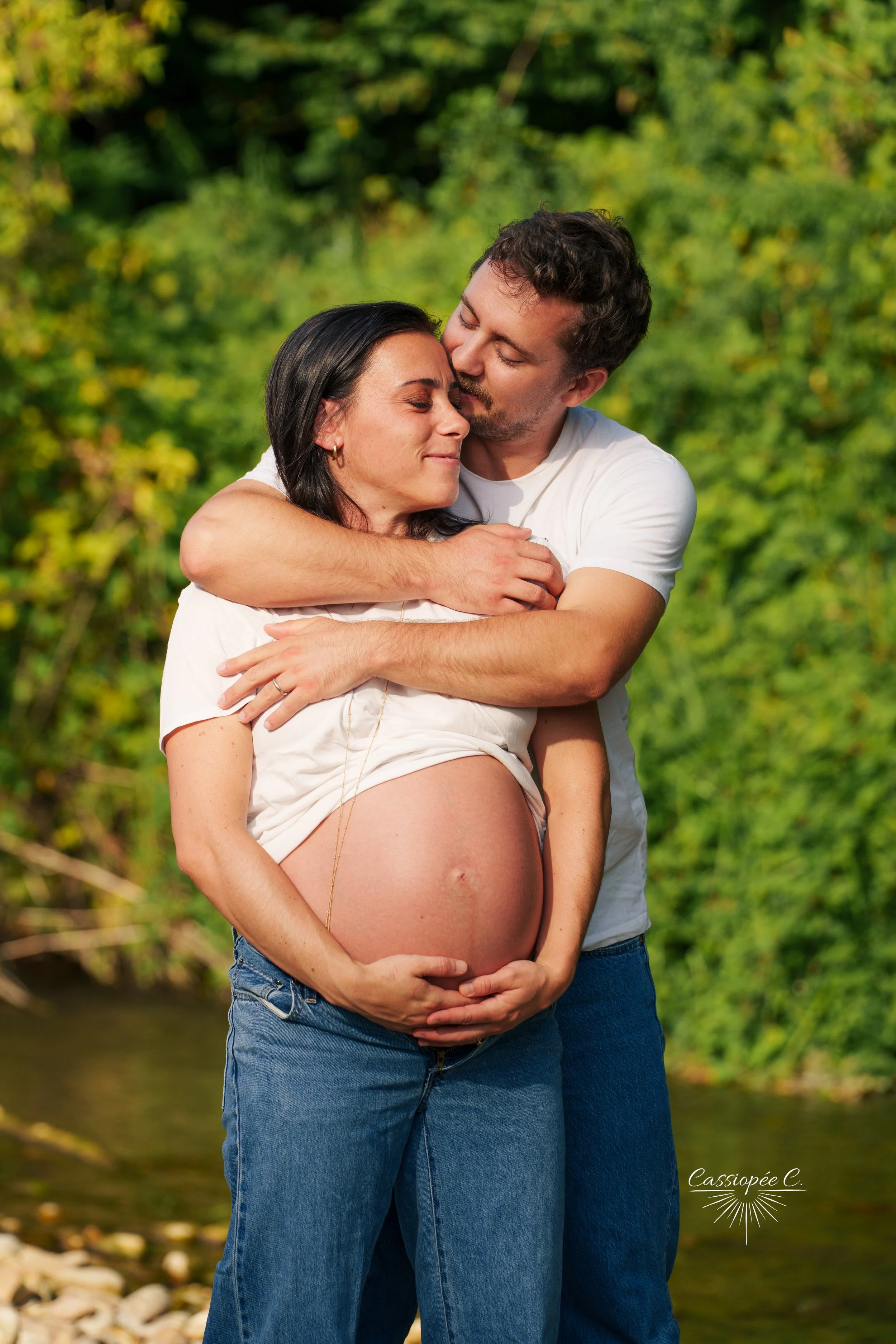 Un couple enceinte se fait un câlin dans la nature, avec un fond de verdure.