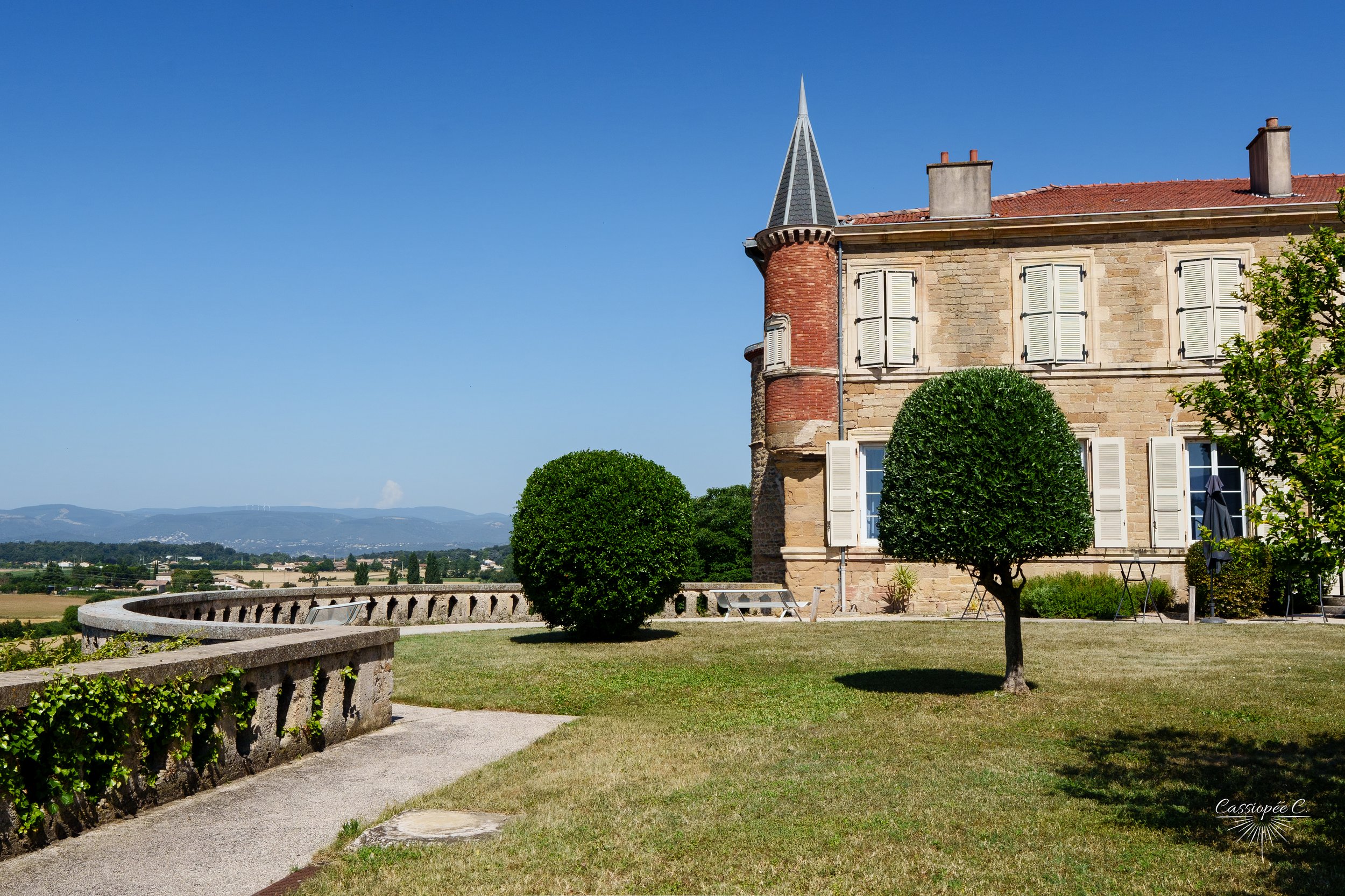 Une maison en pierre avec des fenêtres à volets blancs, un toit en tuiles rouges et une tourelle avec une toiture pointue, entourée d'arbres et d'un jardin bien entretenu, avec un ciel bleu clair en arrière-plan.