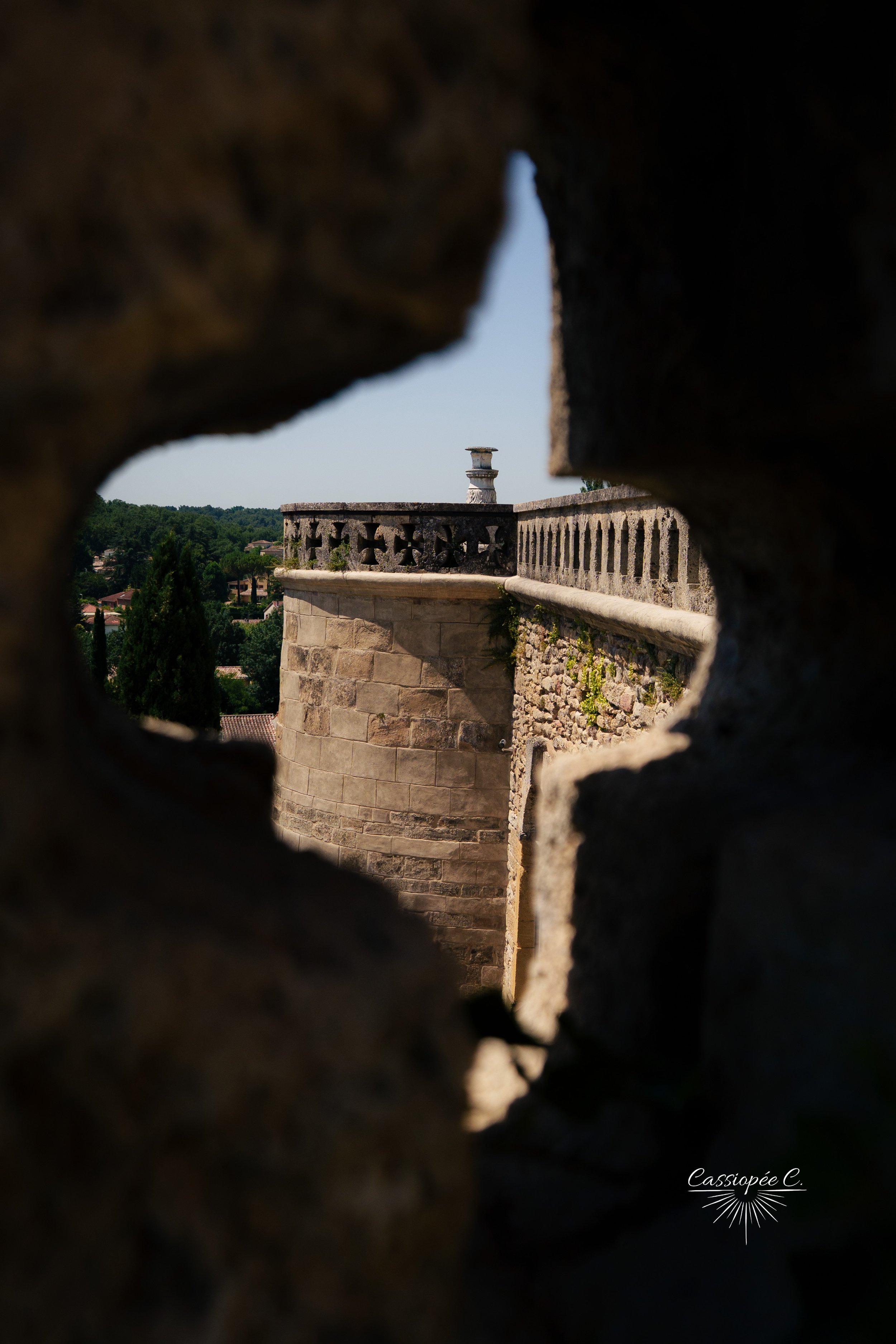 Une vue d'une tour en pierre entourée de remparts, prise à travers une ouverture dans un mur en pierre, avec un paysage verdoyant en arrière-plan.