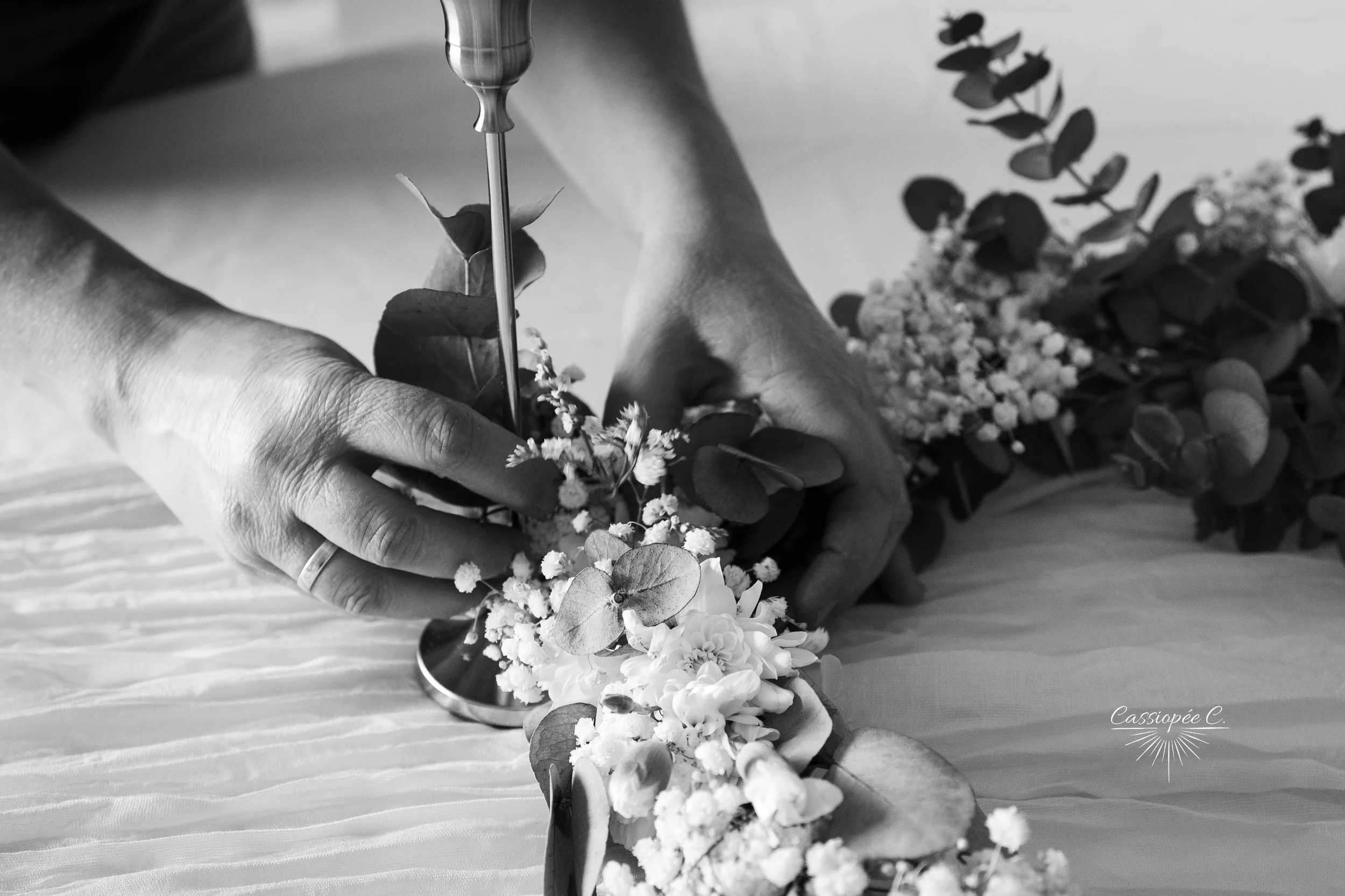 Mains manipulant des fleurs et des feuilles, en train de réaliser une composition florale, avec un fond flou montrant des fleurs et des feuilles.