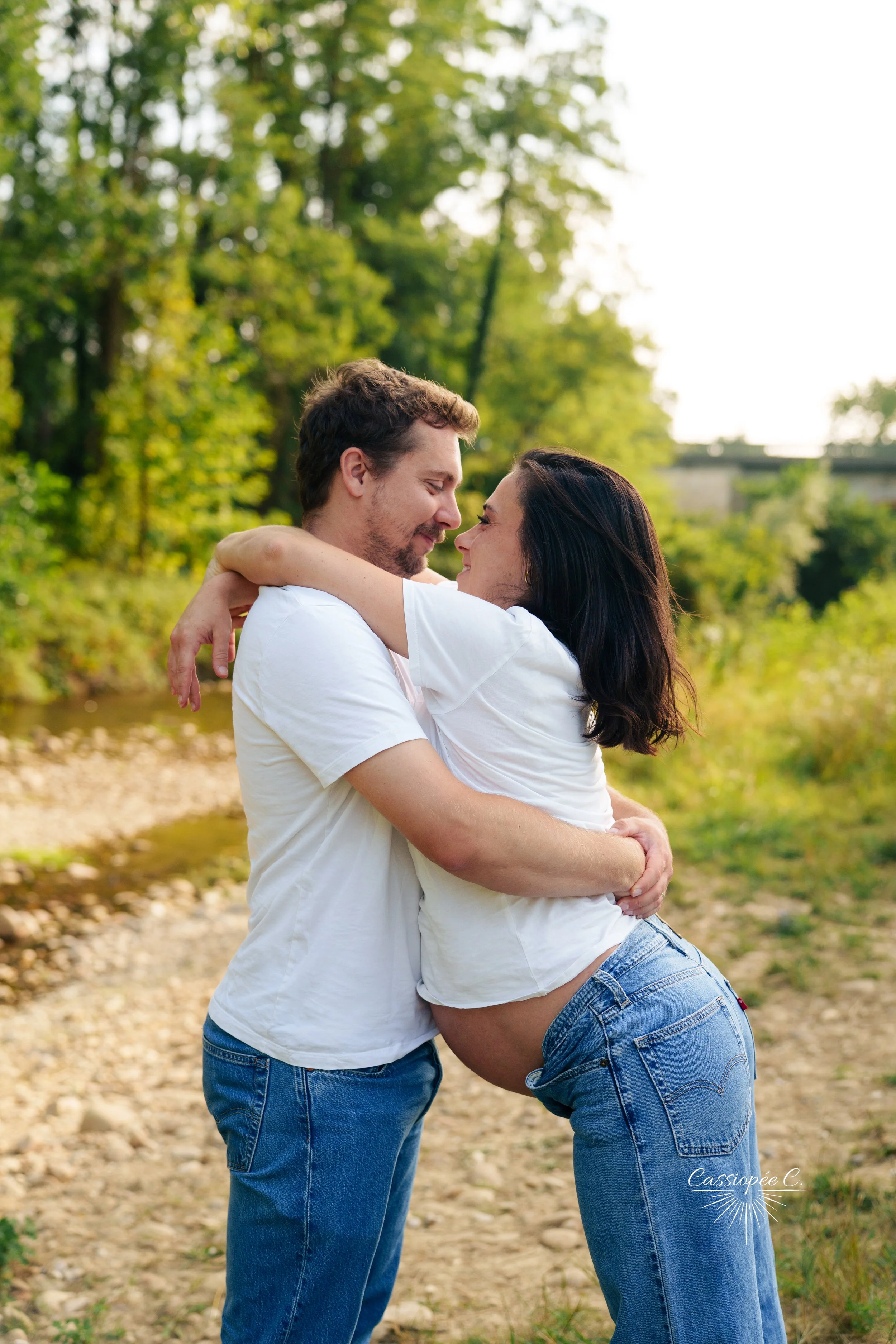 Un couple s'embrasse tendrement dans un cadre naturel avec des arbres et une rivière, ils sourient et semblent heureux.