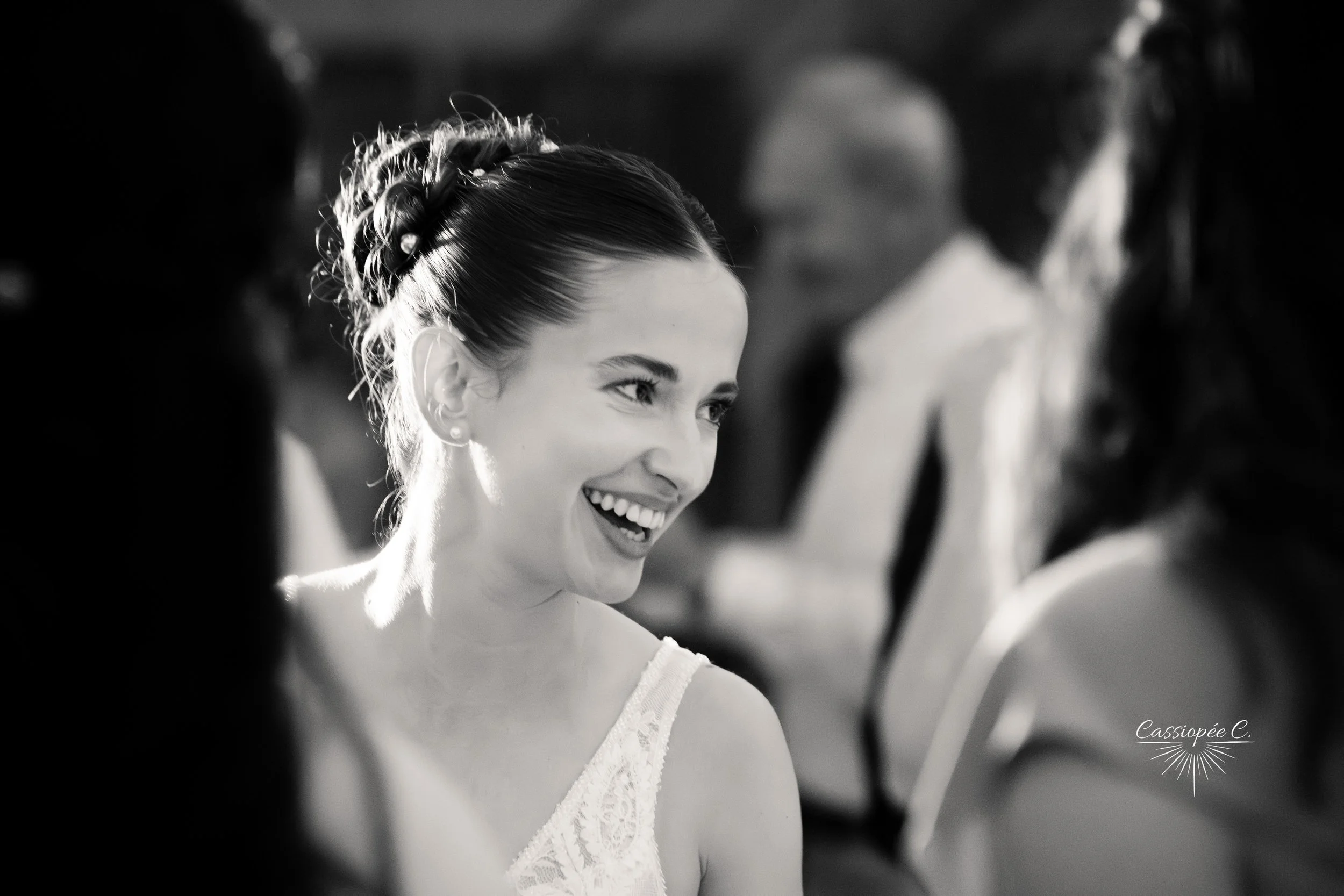 Une femme souriante avec une coiffure élaborée, portant une robe en dentelle, entourée de plusieurs autres personnes dans un cadre probablement festif ou cérémonial.