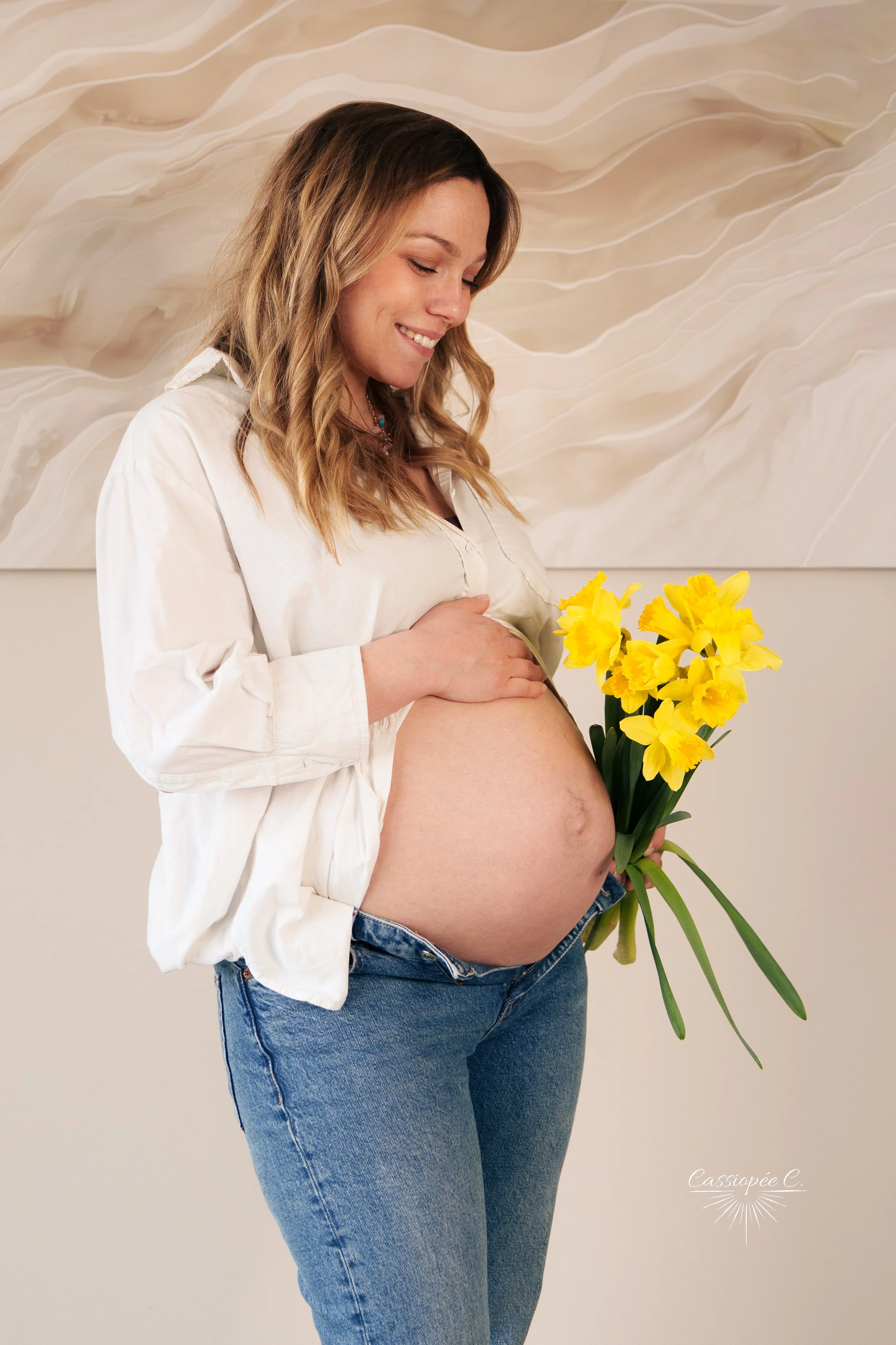 Femme enceinte tenant des fleurs jaunes et portant une chemise blanche avec un fond artistique abstrait.