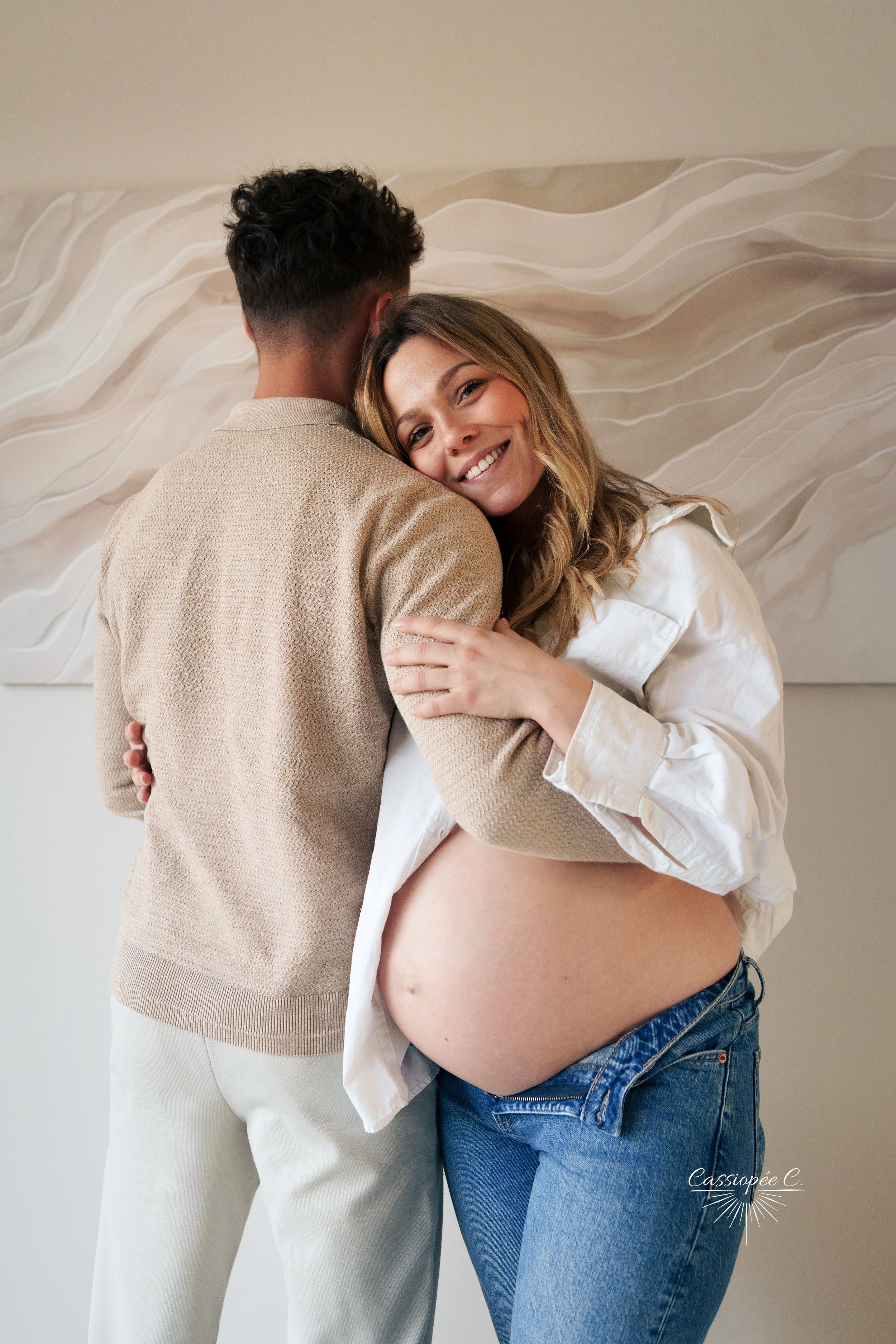 Une femme enceinte souriante, portant un chemisier blanc décolleté et un jean, étant embrassée par un homme dont le dos est visible, portant un pull beige clair, devant un mur blanc avec une œuvre d'art abstraite