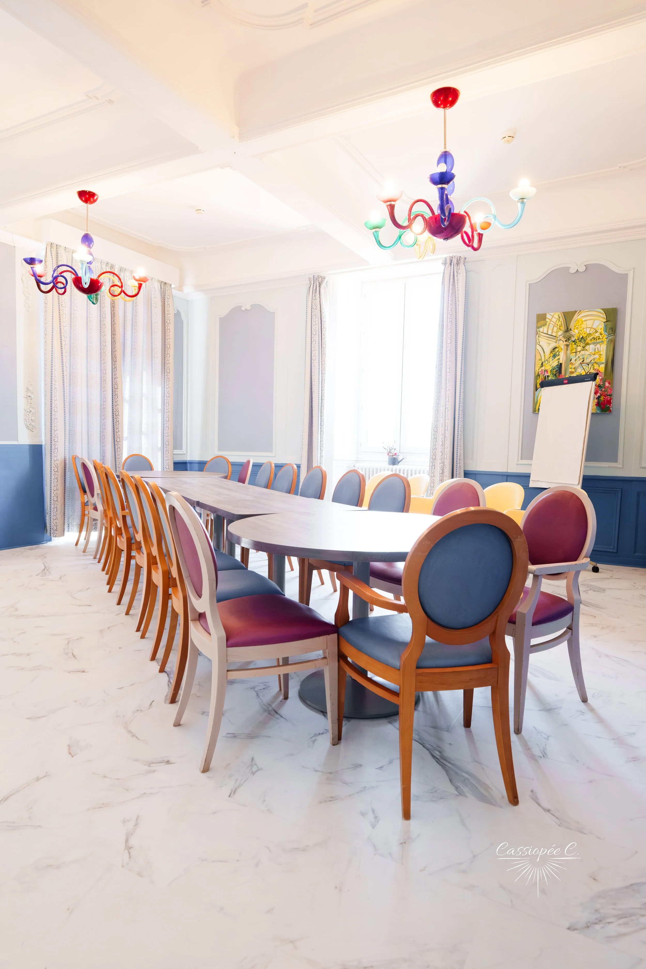 Salle de réunion avec une grande table ovale entourée de chaises aux couleurs pastel, deux lustres colorés au plafond, rideaux blancs, peinture florale sur le mur, sol en marbre blanc, éclairée par une grande fenêtre en arrière-plan.