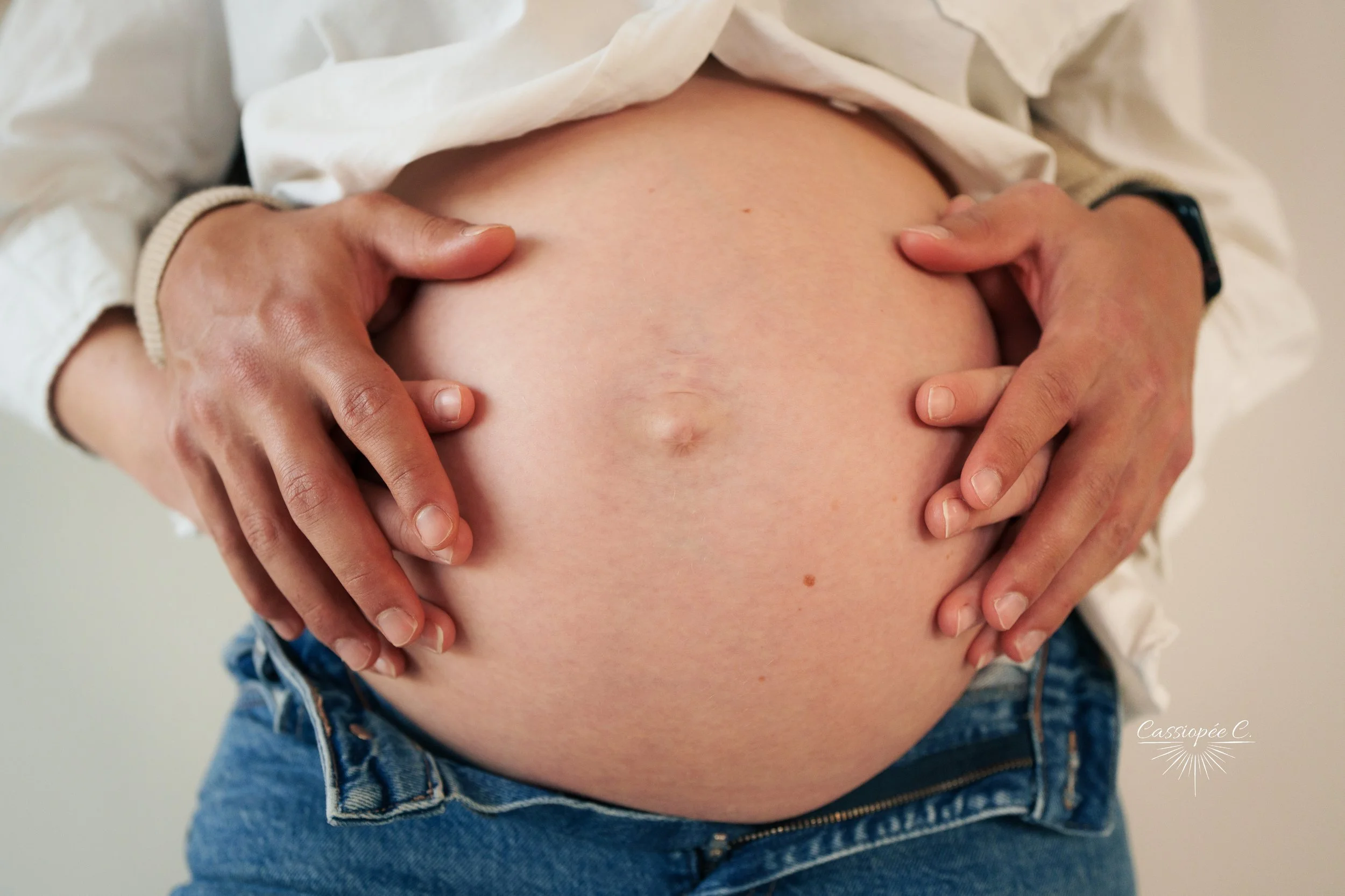 Un ventre de femme enceinte avec quatre mains posées dessus, deux de chaque côté, portant un vêtement blanc et un jean.
