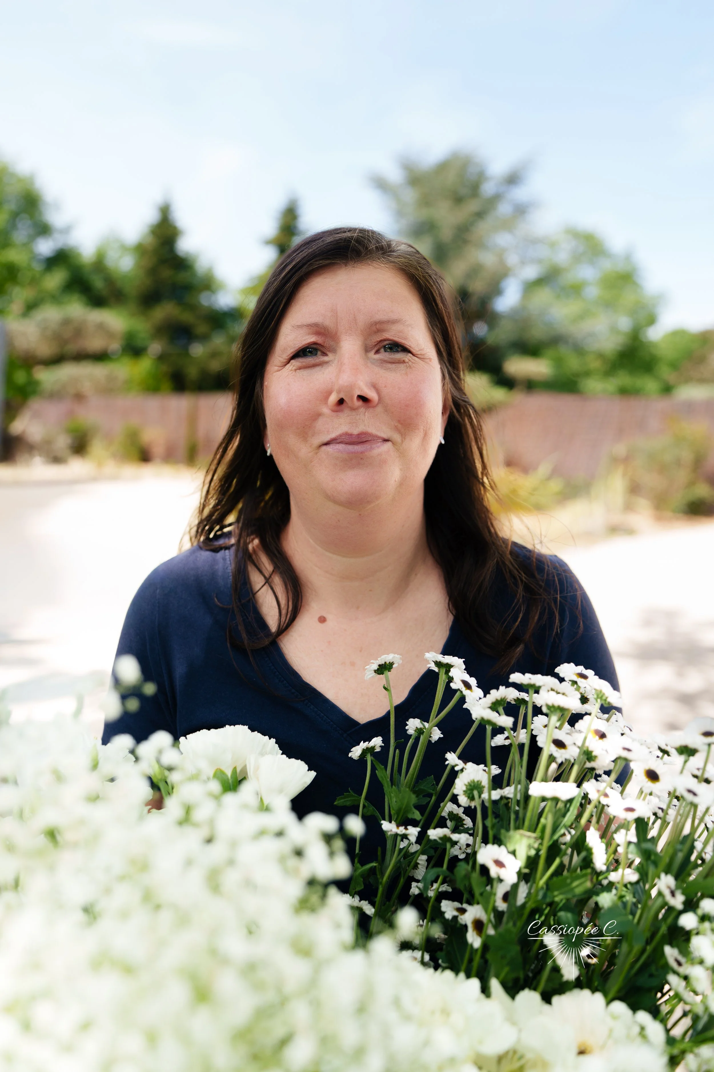Une femme avec des cheveux bruns foncés, portant un haut bleu, se trouve devant un fond de jardin avec des arbres et un mur en brique. Elle se tient derrière un bouquet de fleurs blanches, notamment des marguerites. C'est une photo prise en extérieur