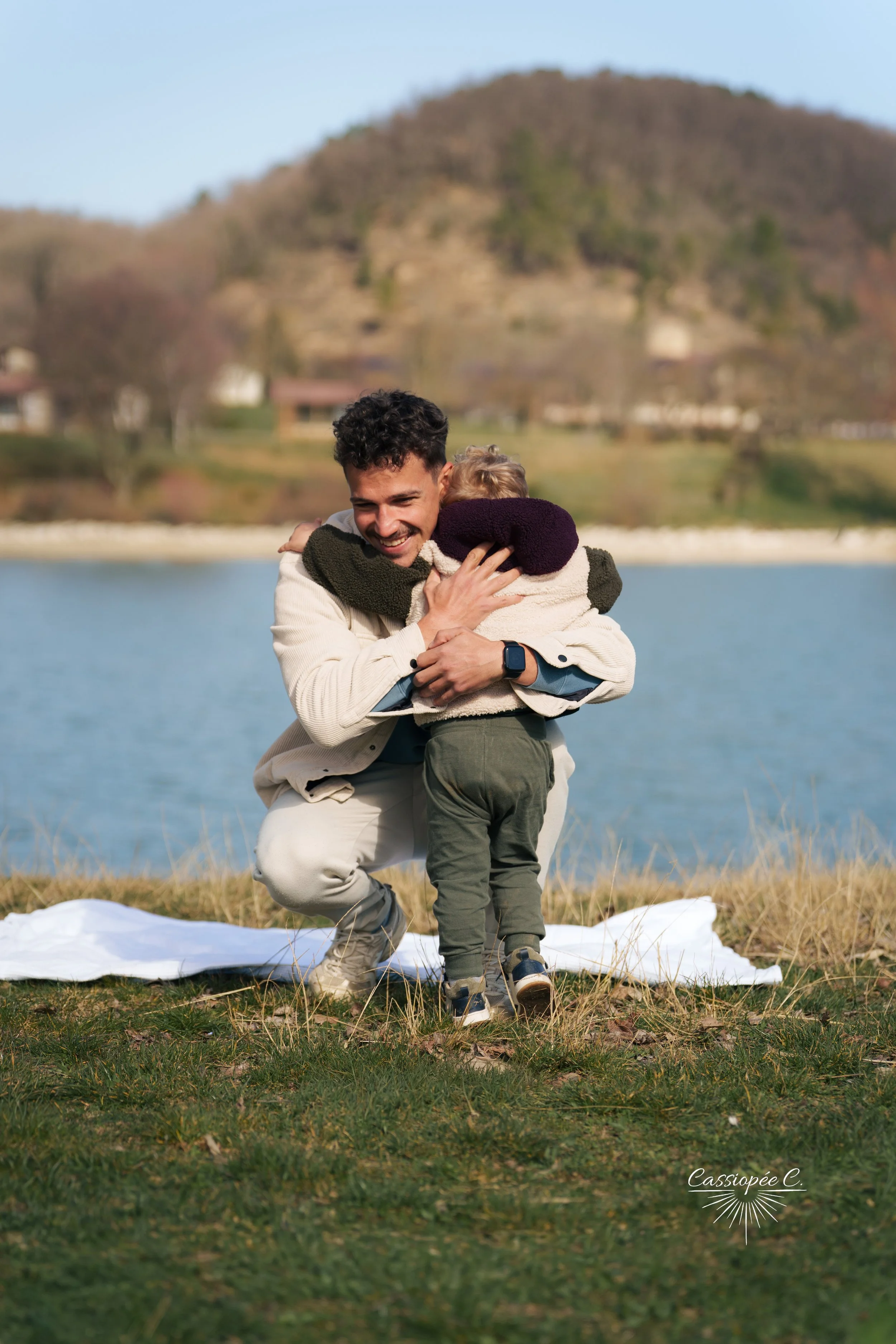 Un homme souriant en tenue beige et son fils, en train de s'étreindre près d'un lac avec un fond de colline, en plein air, lors d'une journée ensoleillée.