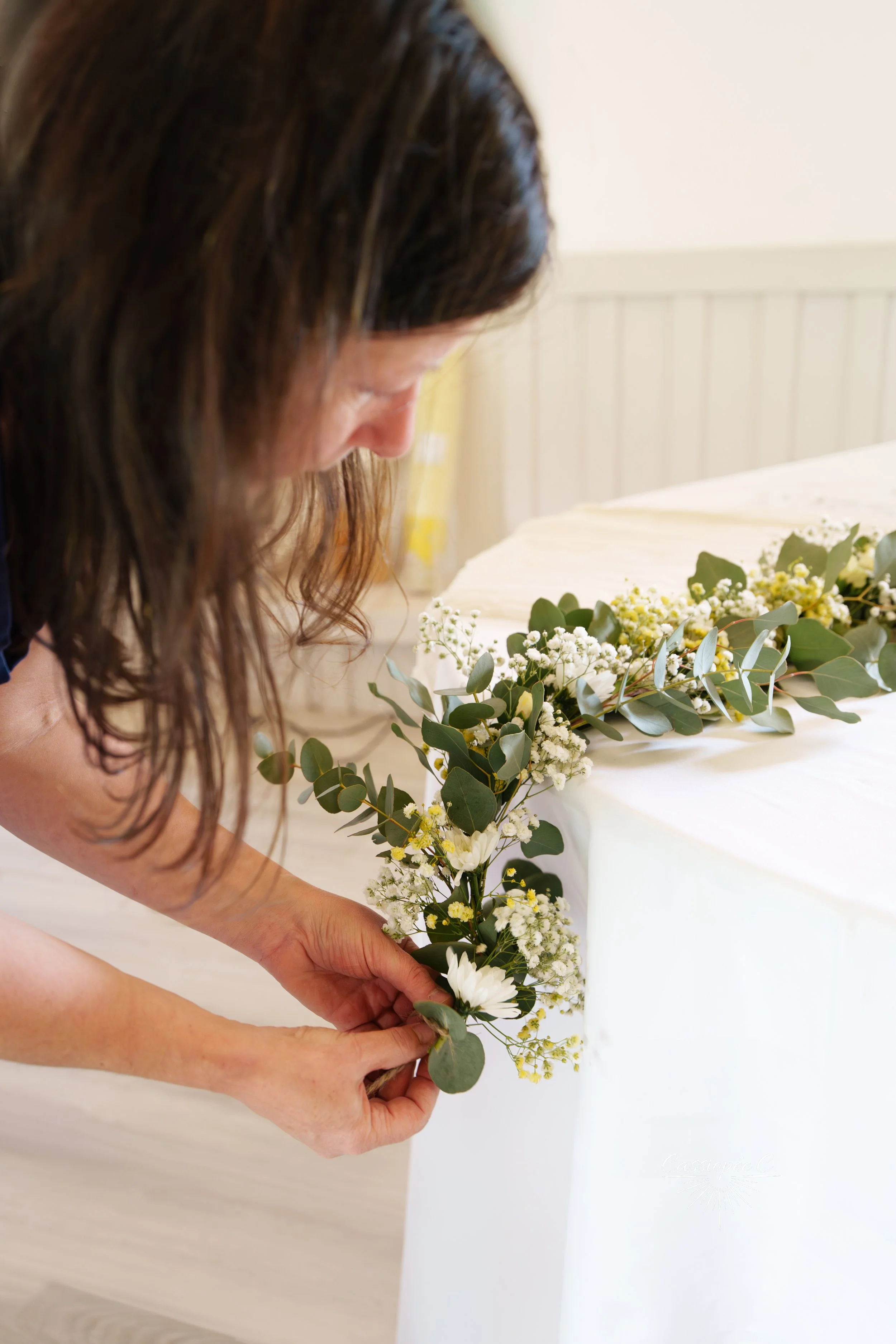 Femme arrangeant un bouquet de fleurs blanches et vertes sur une table, fond blanc avec pan de mur en bois clair.