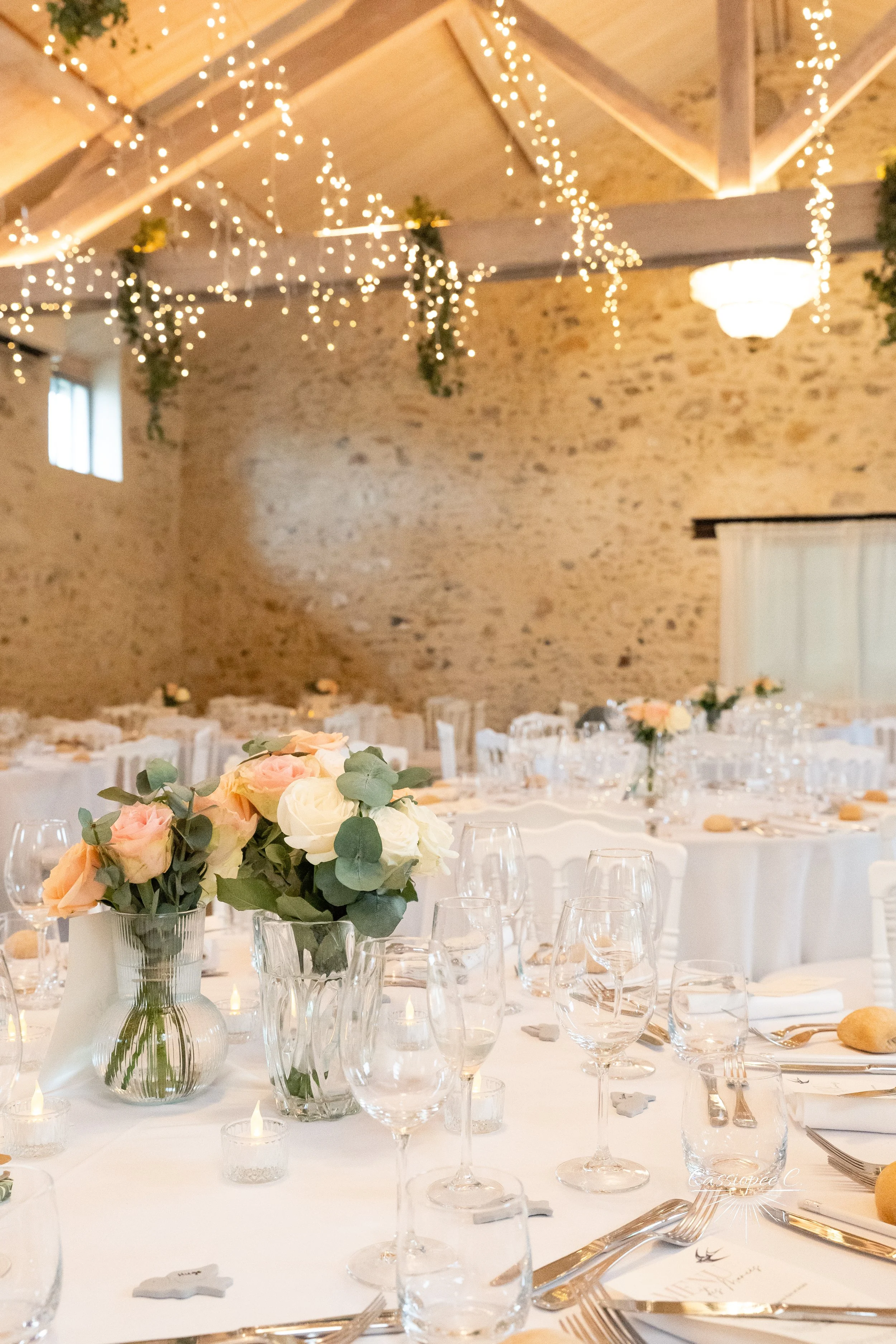Décor de mariage avec tables ovales, centres de fleurs roses et blanches, bougies, et guirlandes lumineuses suspendues sous un plafond en bois dans une salle rustique en pierre.