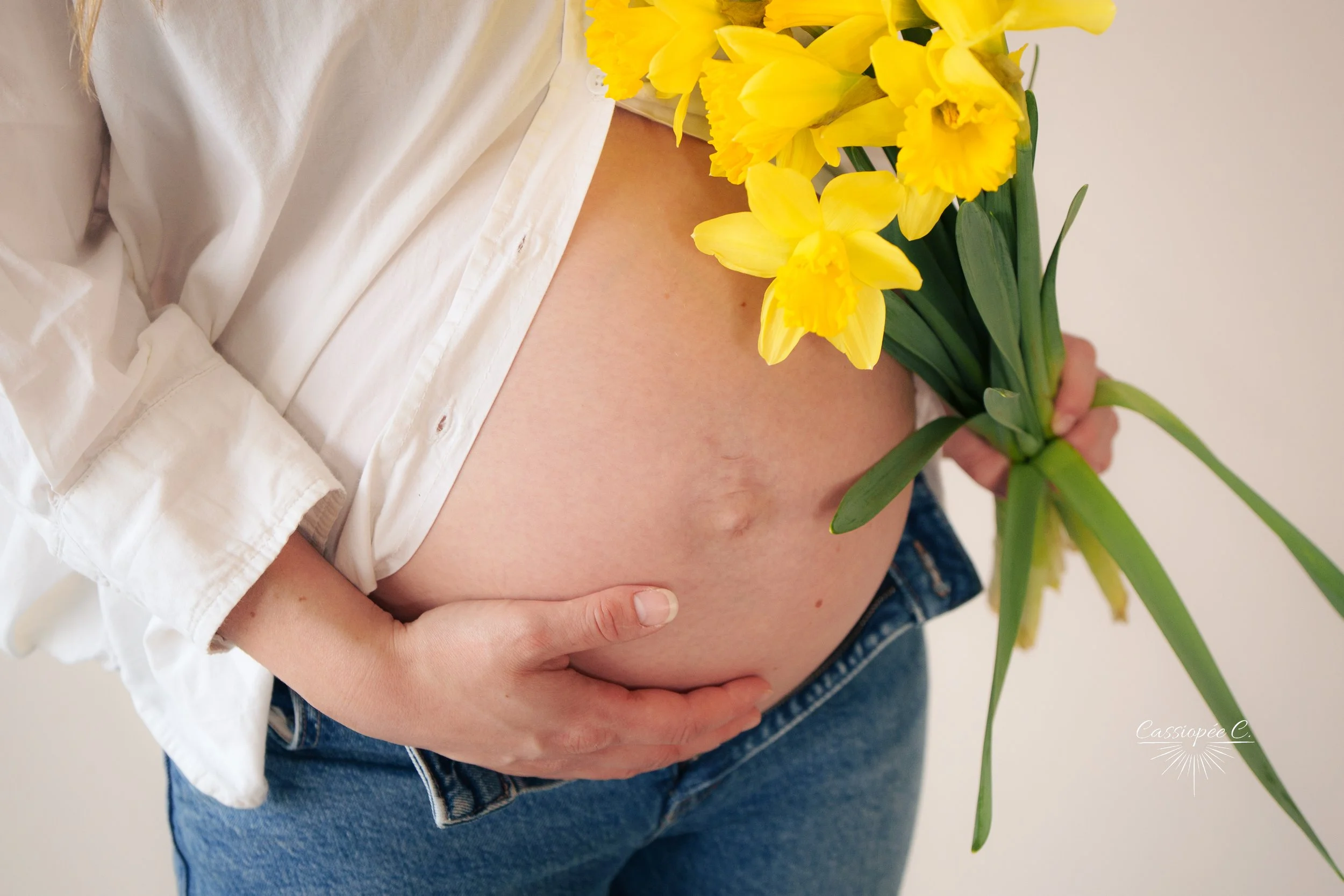 Une femme enceinte tient un bouquet de jonquilles jaunes, portant une chemise blanche partiellement ouverte et un jean bleu.