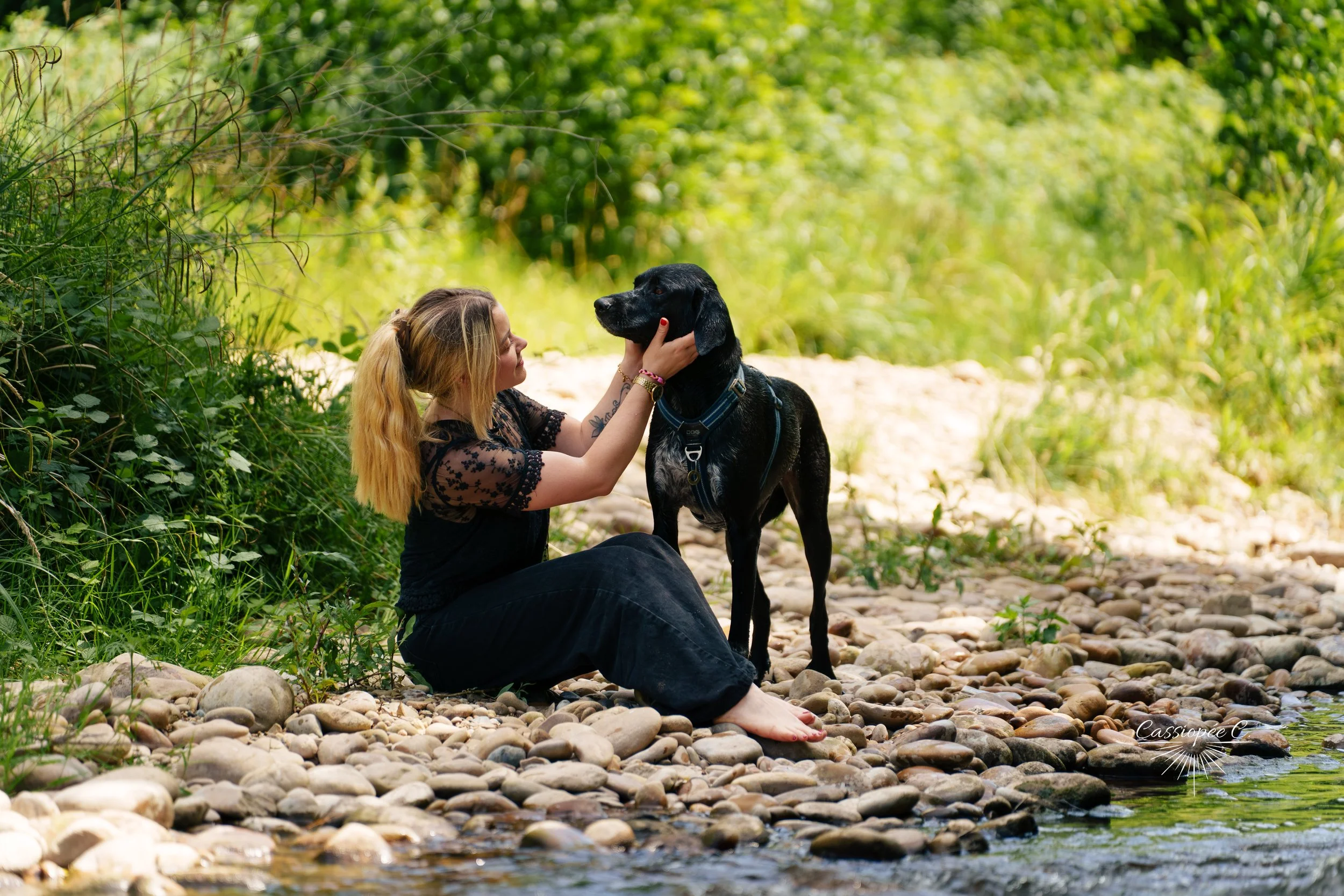 Une femme aux cheveux blonds assise au bord d'une rivière, caressant un chien noir. La scène se déroule dans une forêt verdoyante.