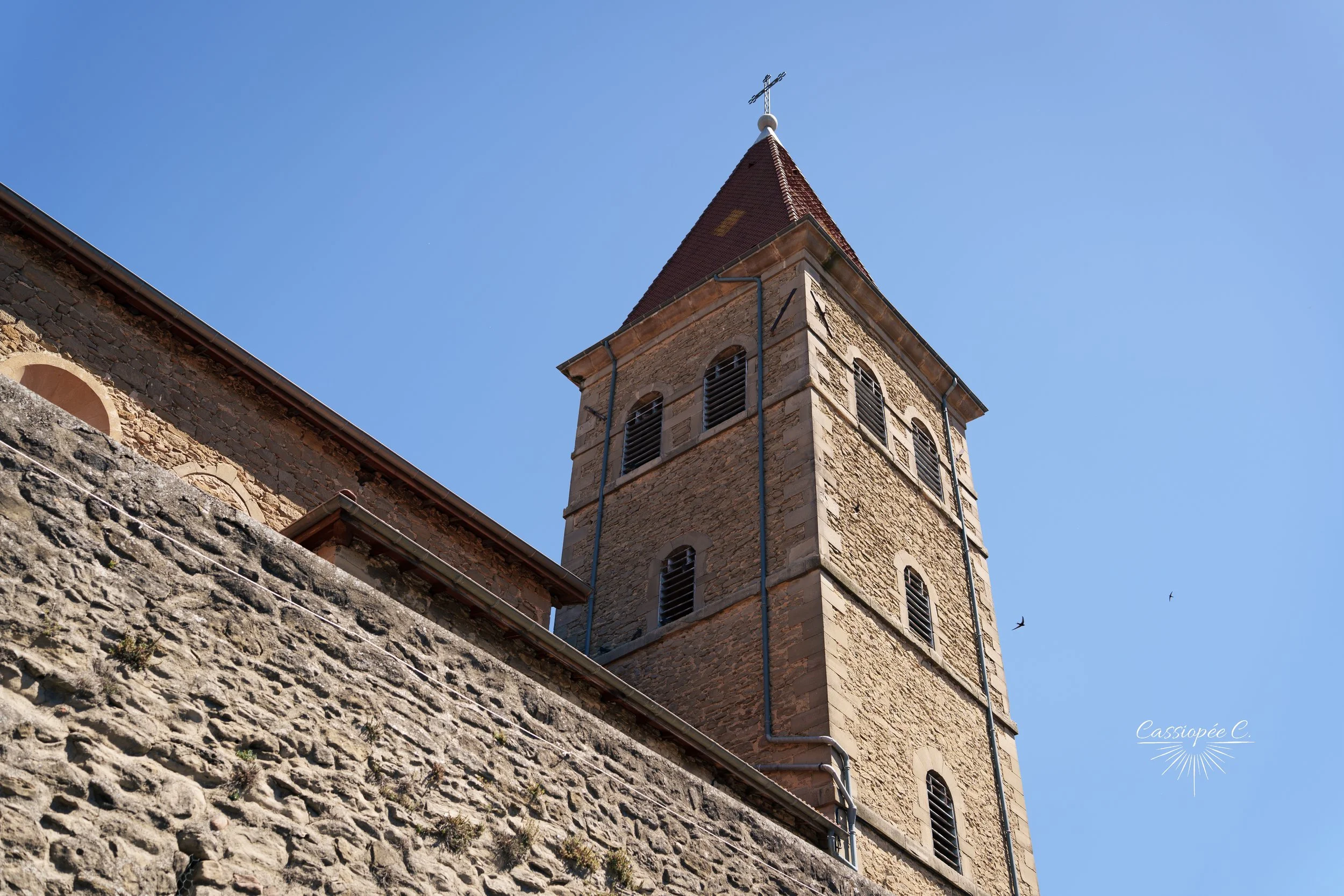 Vue d'une tour d'une vieille église en pierre avec un toit en pente, un croix au sommet, et un mur en pierre en premier plan, sous un ciel clair.
