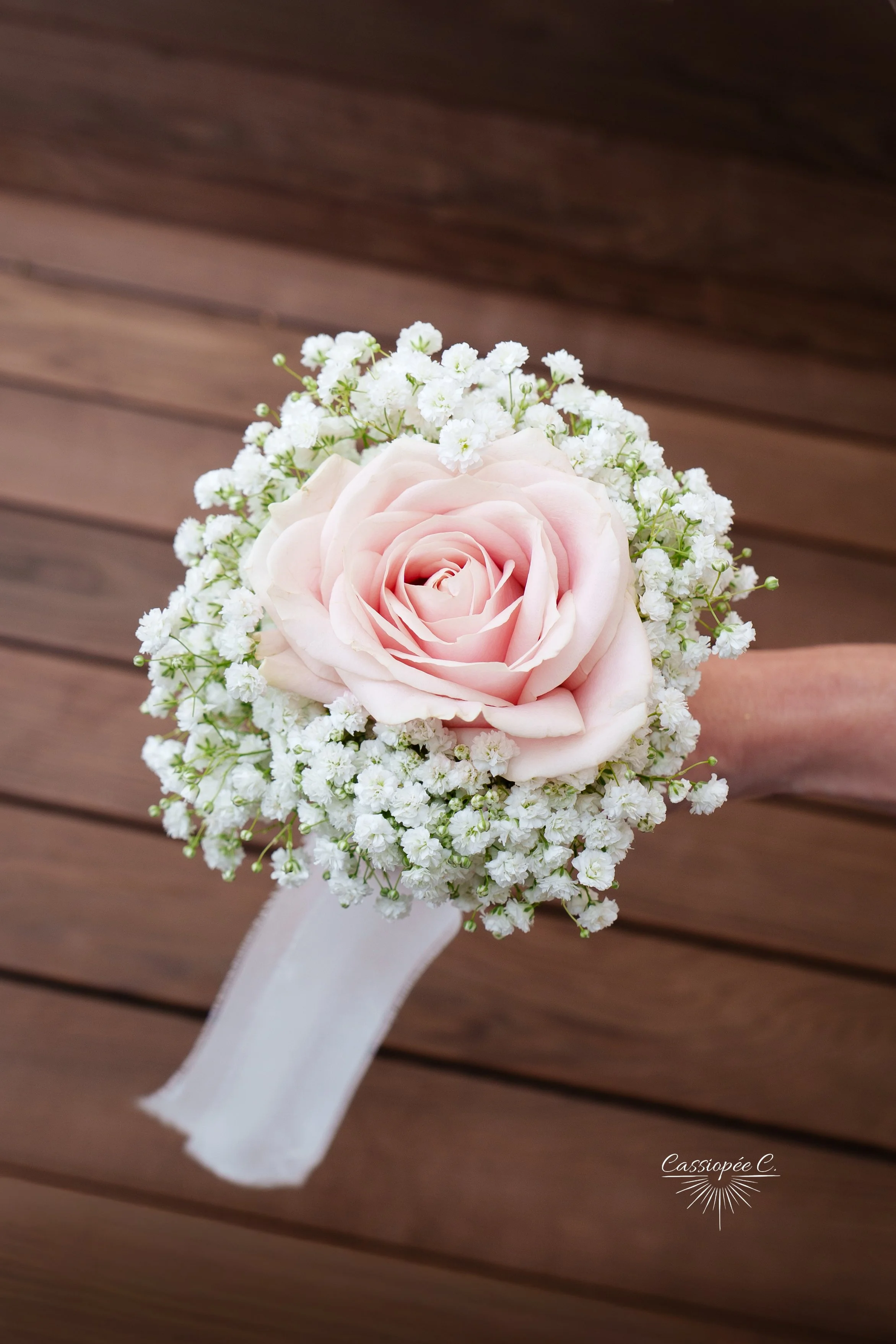 Un bouquet de fleurs composé d'une grande rose rose pâle entourée de petites fleurs blanches, attaché avec un ruban blanc, contre un fond en bois.