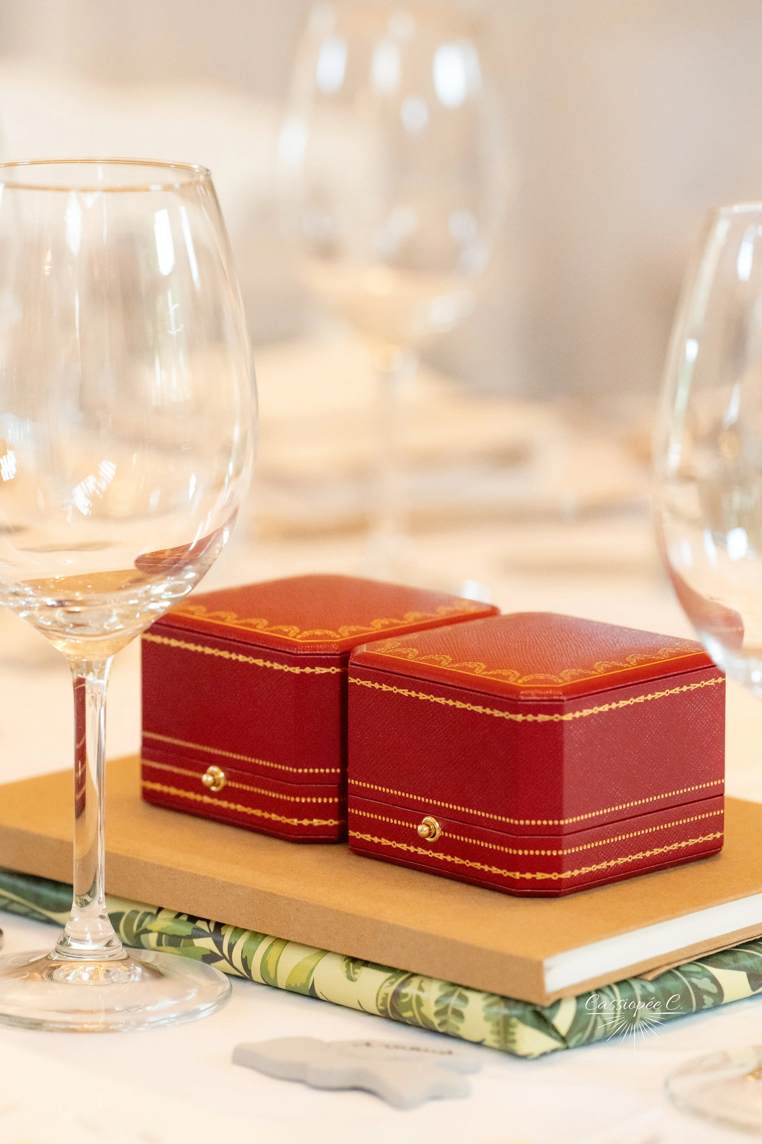 Deux boîtes rouges à alliances avec des motifs dorés, placées sur un plateau en bois, entourées de verres à vin vides et de carnets, dans un cadre élégant et bien éclairé.