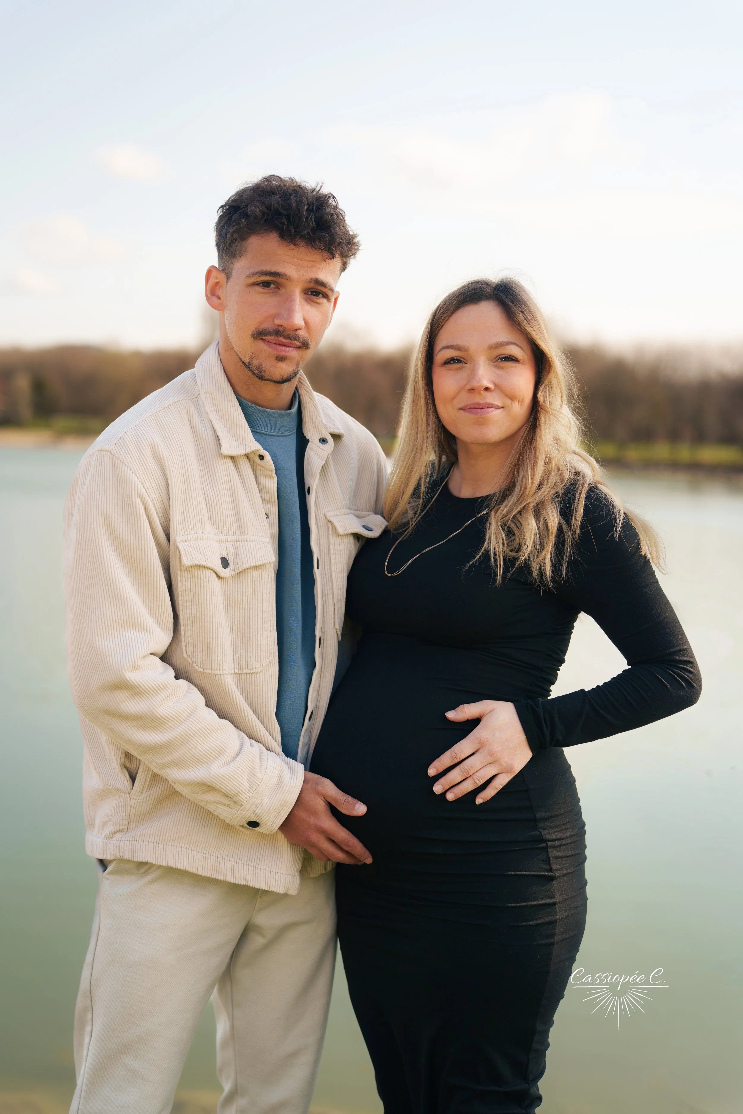 Un couple posant devant un lac avec des arbres en arrière-plan, la femme enceinte elle tient son ventre avec une main et l'homme a une main sur la taille de la femme.