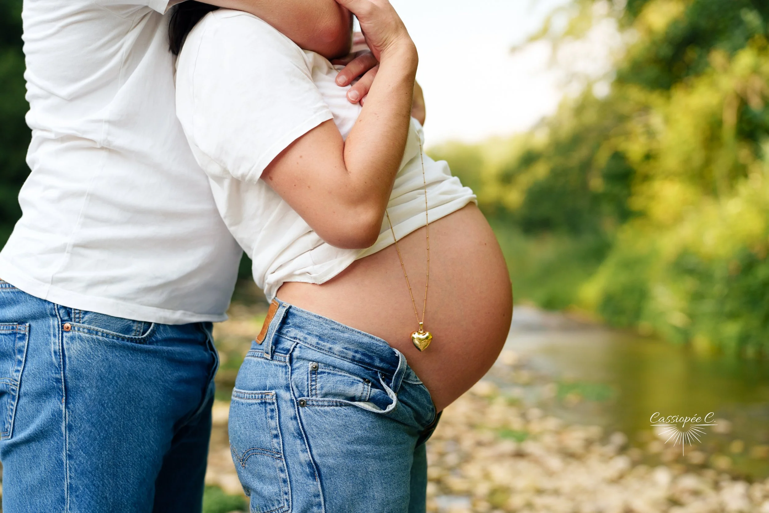 Une future maman portant un t-shirt blanc et un jeans, avec un ventre rond, à côté d'une personne portant également un jeans, dans un champ en plein air, avec beaucoup de verdure.