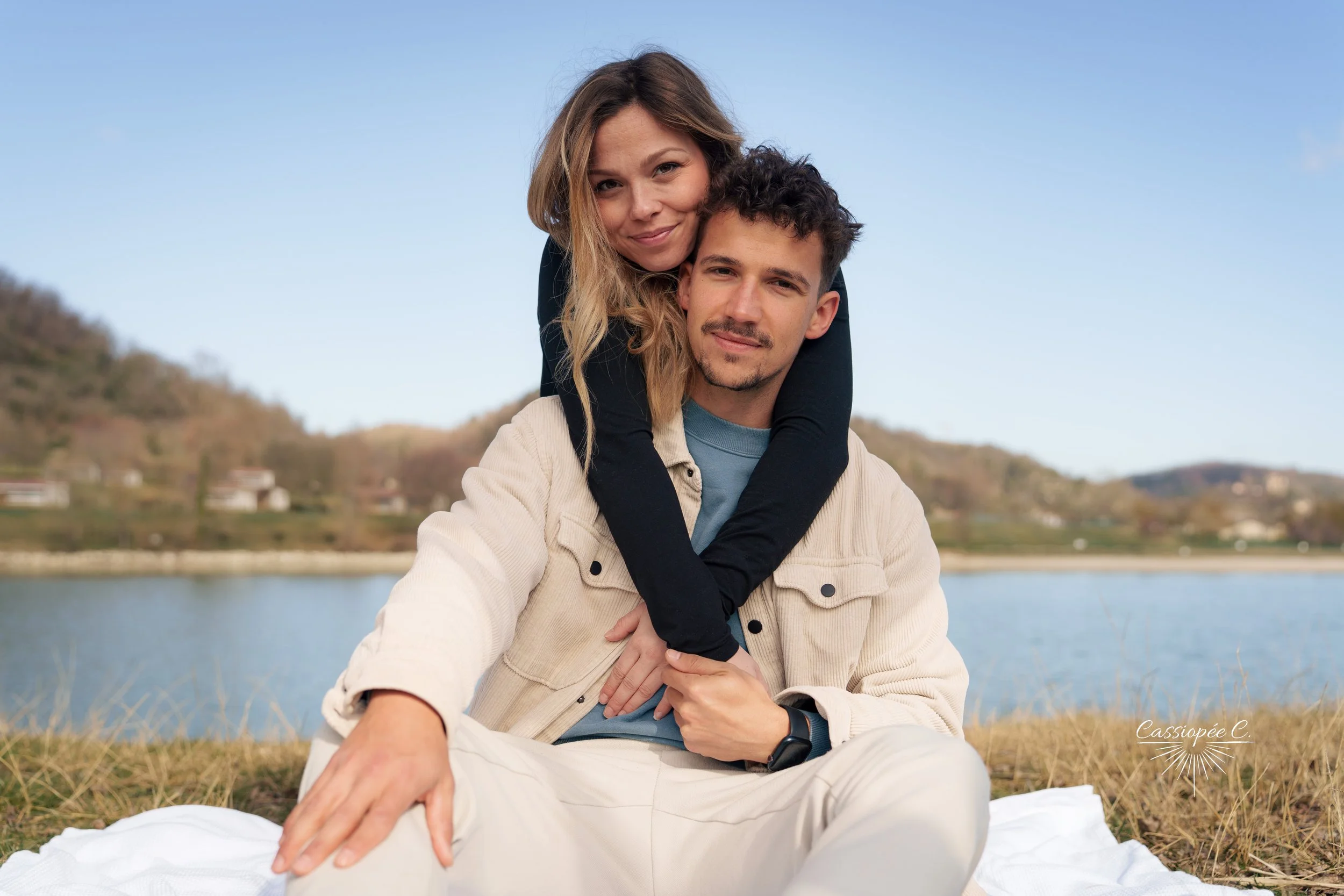 Un couple assis près d'un lac avec des collines verdoyantes en arrière-plan, la femme est sur le dos de l'homme avec un sourire, le paysage est paisible et ensoleillé.
