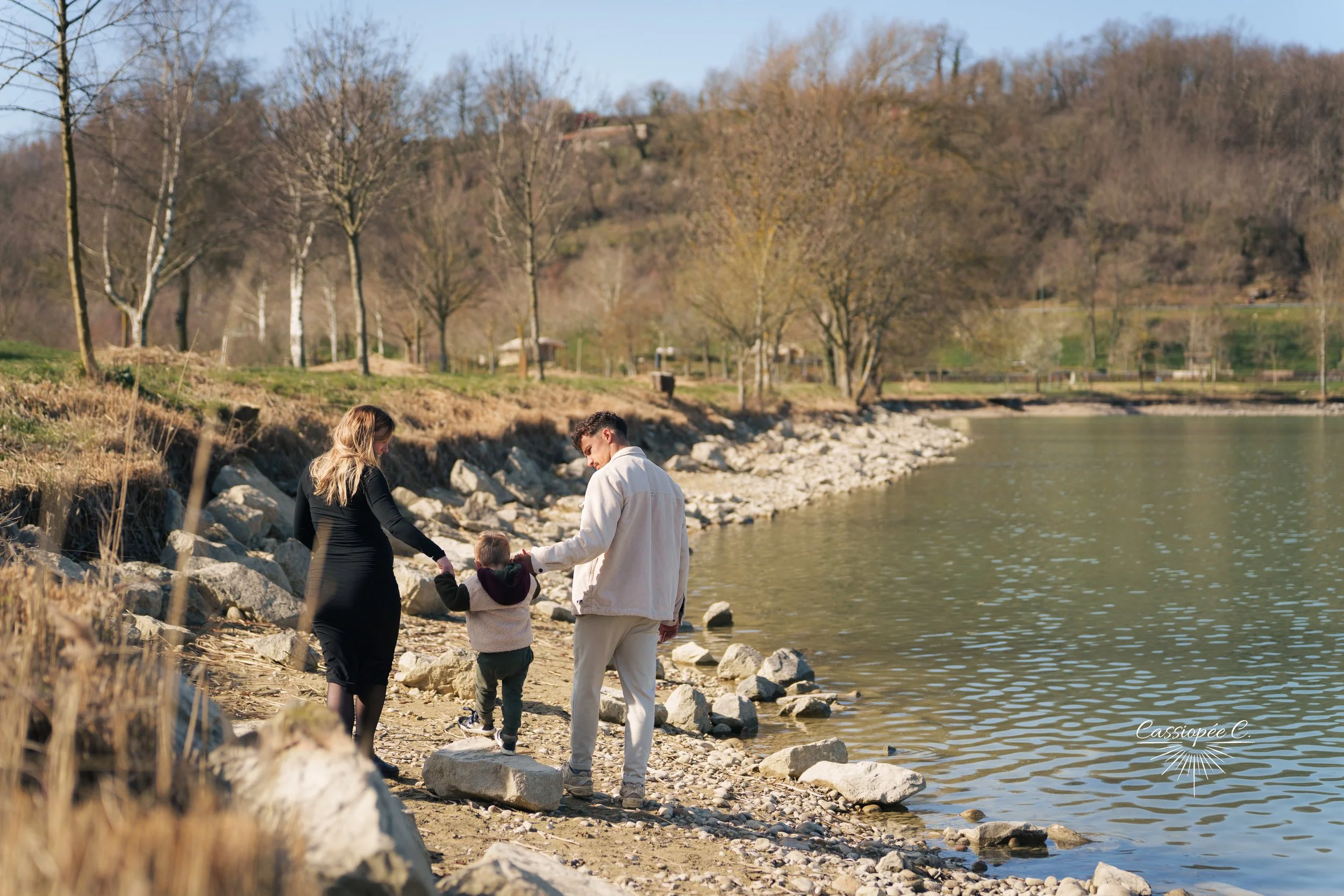 Un groupe de trois personnes, deux adultes et un enfant, marche le long du rivage d'une rivière. Le paysage est entouré d'arbres sans feuilles et de collines en arrière-plan, dans une atmosphère calme et ensoleillée.
