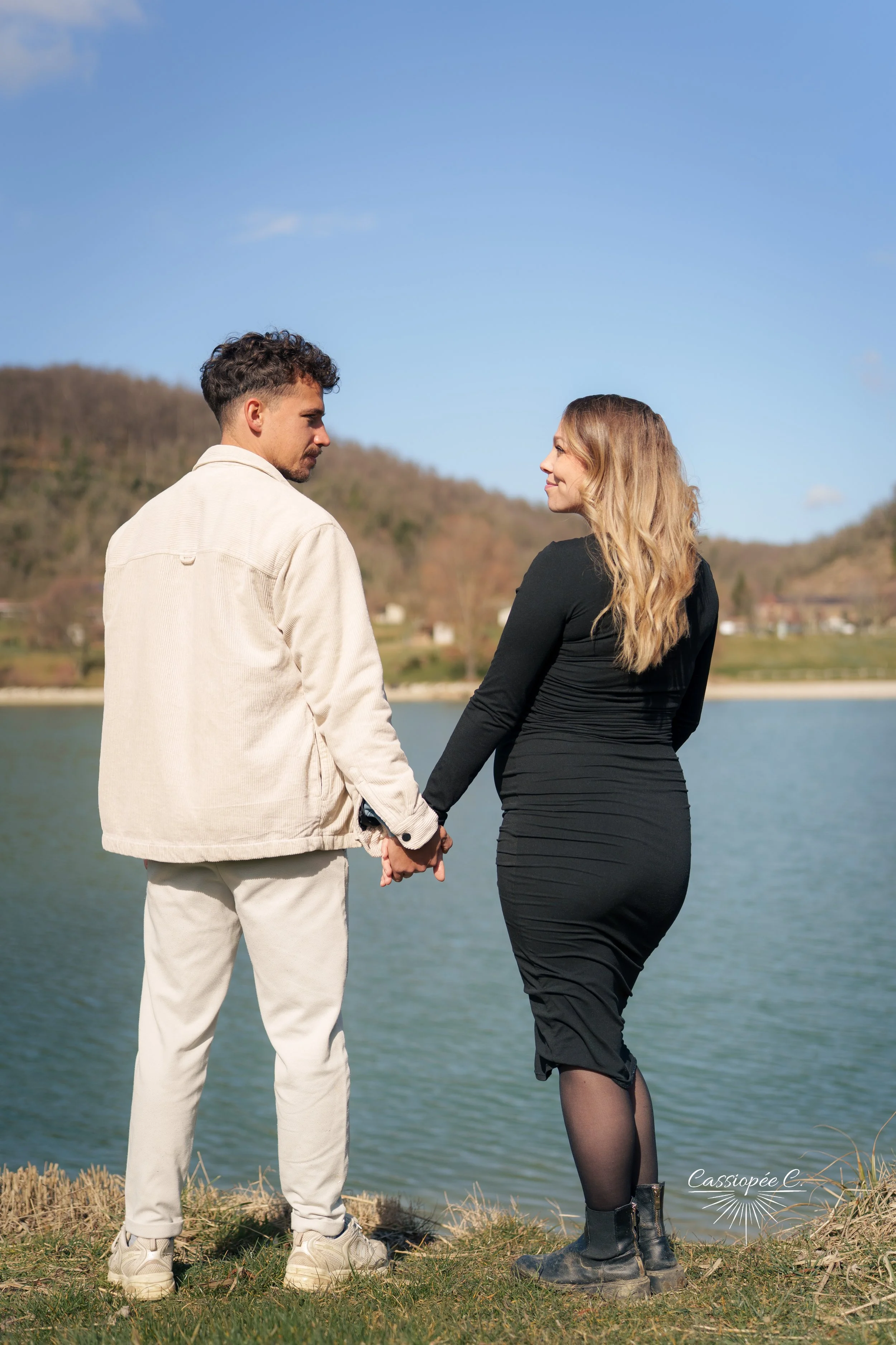 Un couple se tient par la main près d'un lac, regardant l'un l'autre, avec une forêt en arrière-plan. Il fait beau avec un ciel bleu clair.