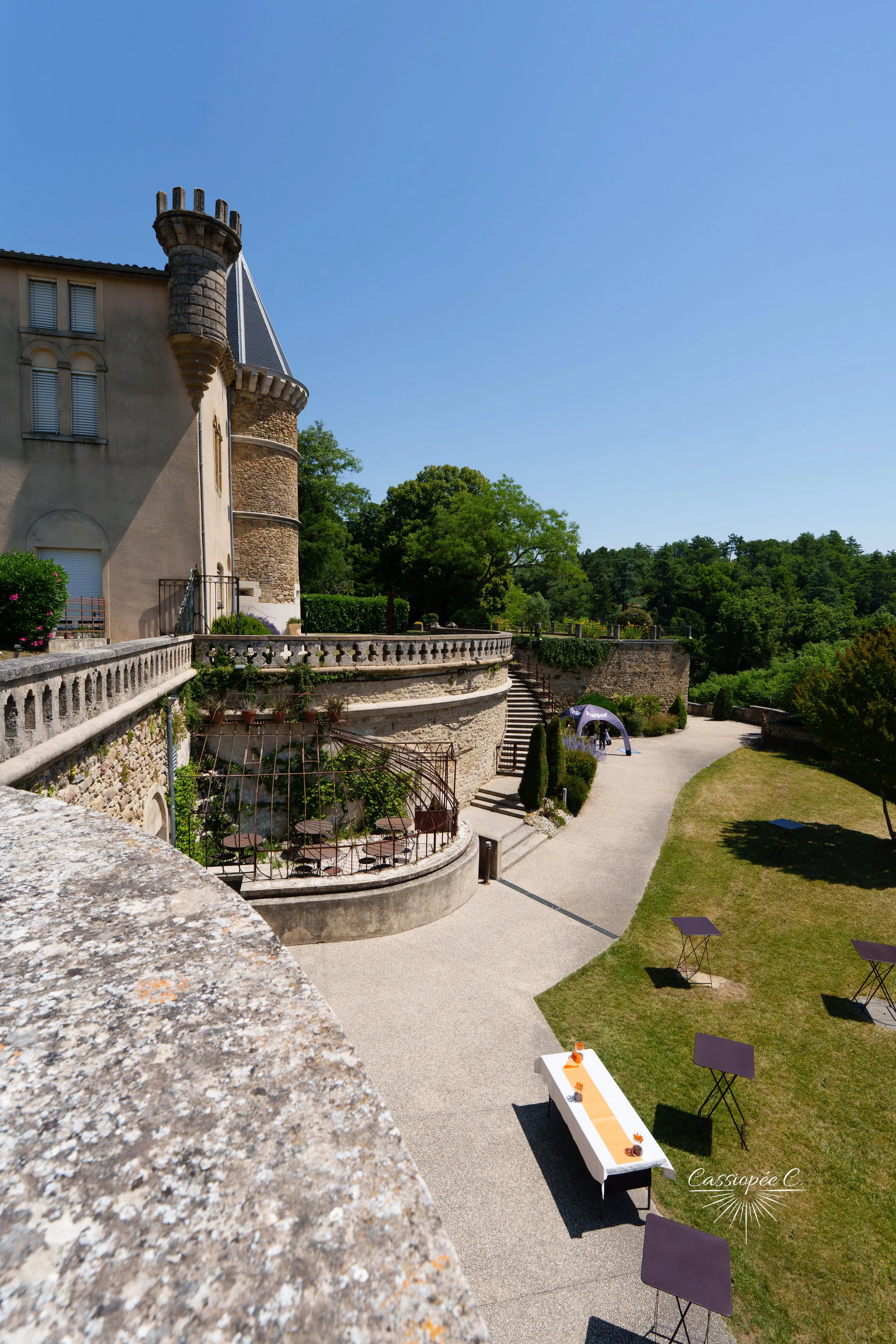 Une scène extérieure d'une terrasse d'un château avec des escaliers en pierre, des tables, un chemin de table orange et des décorations, dans un environnement verdoyant et ensoleillé.