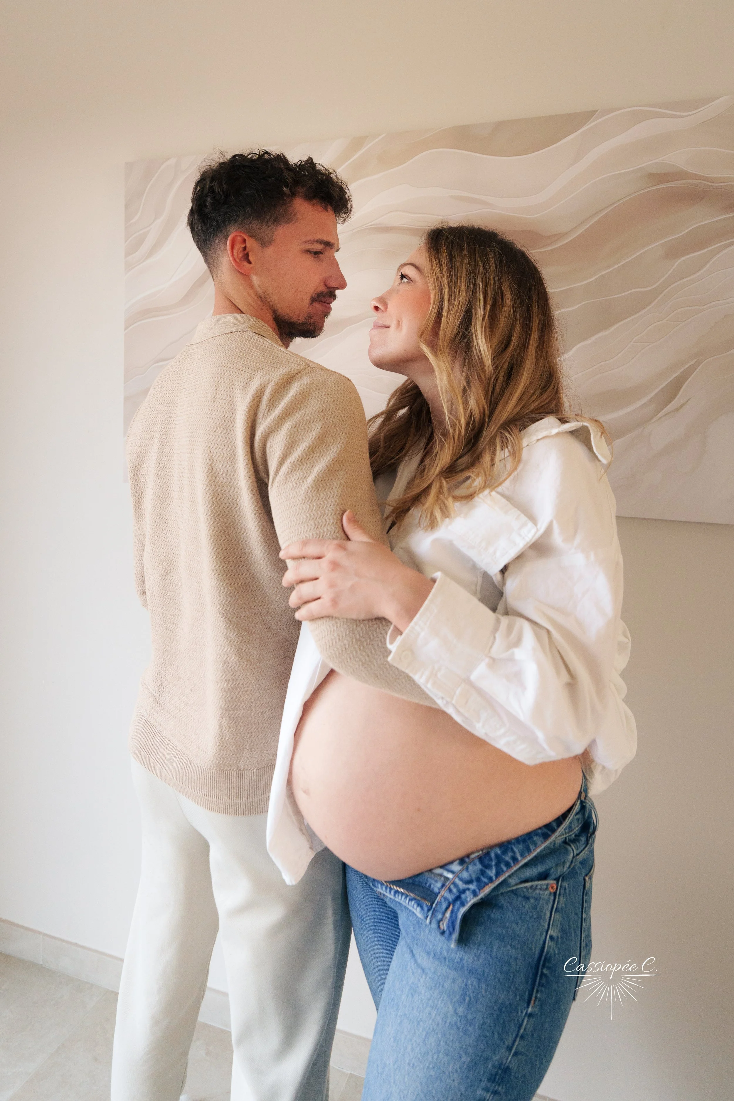 Une femme enceinte et un homme se regardant tendrement, la femme tenant le ventre arrondi de sa grossesse, dans une pièce avec une œuvre d'art abstraite en fond.