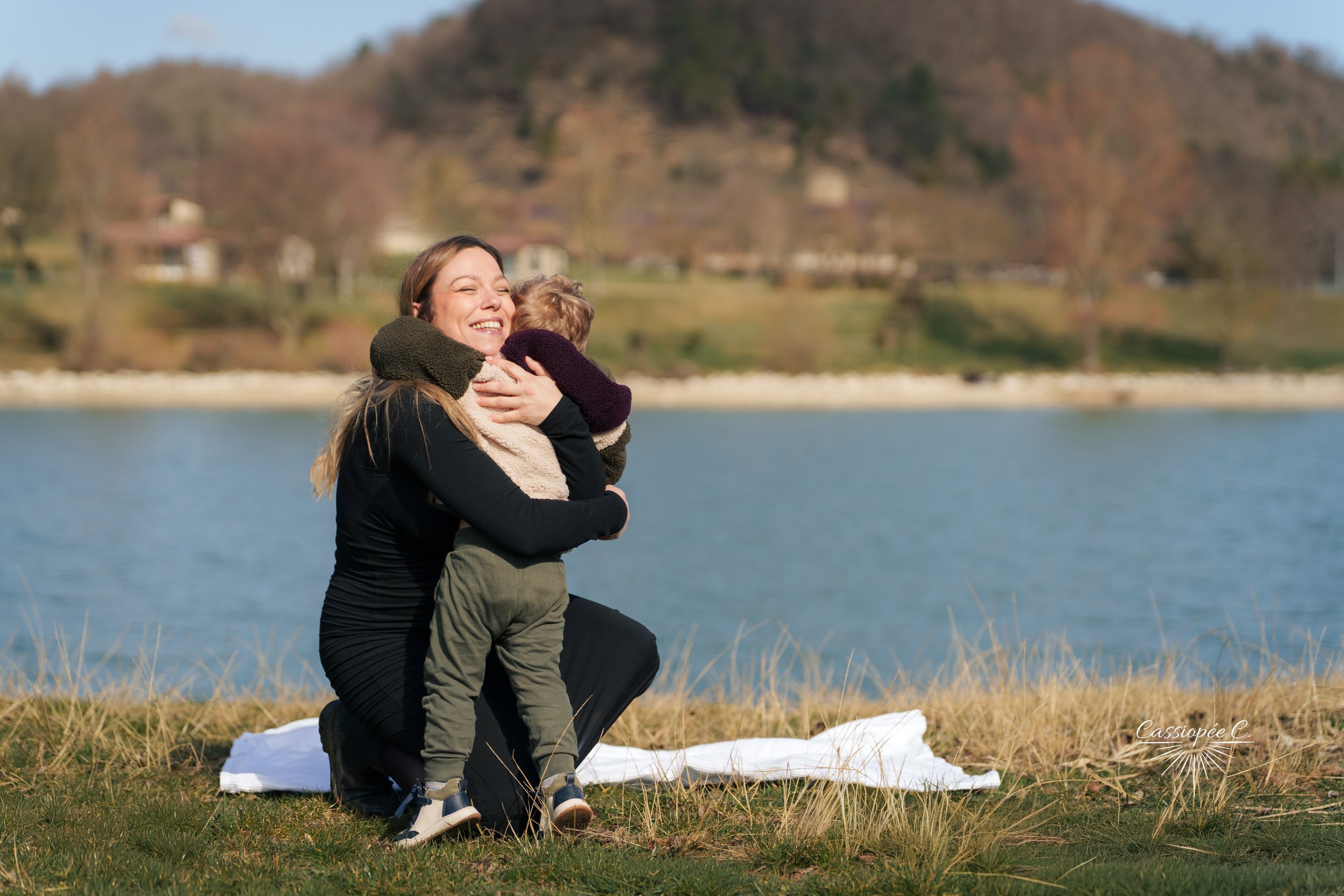 Une femme souriante et un jeune garçon dans un paysage au bord d'un lac, avec des montagnes en arrière-plan, en pleine journée.