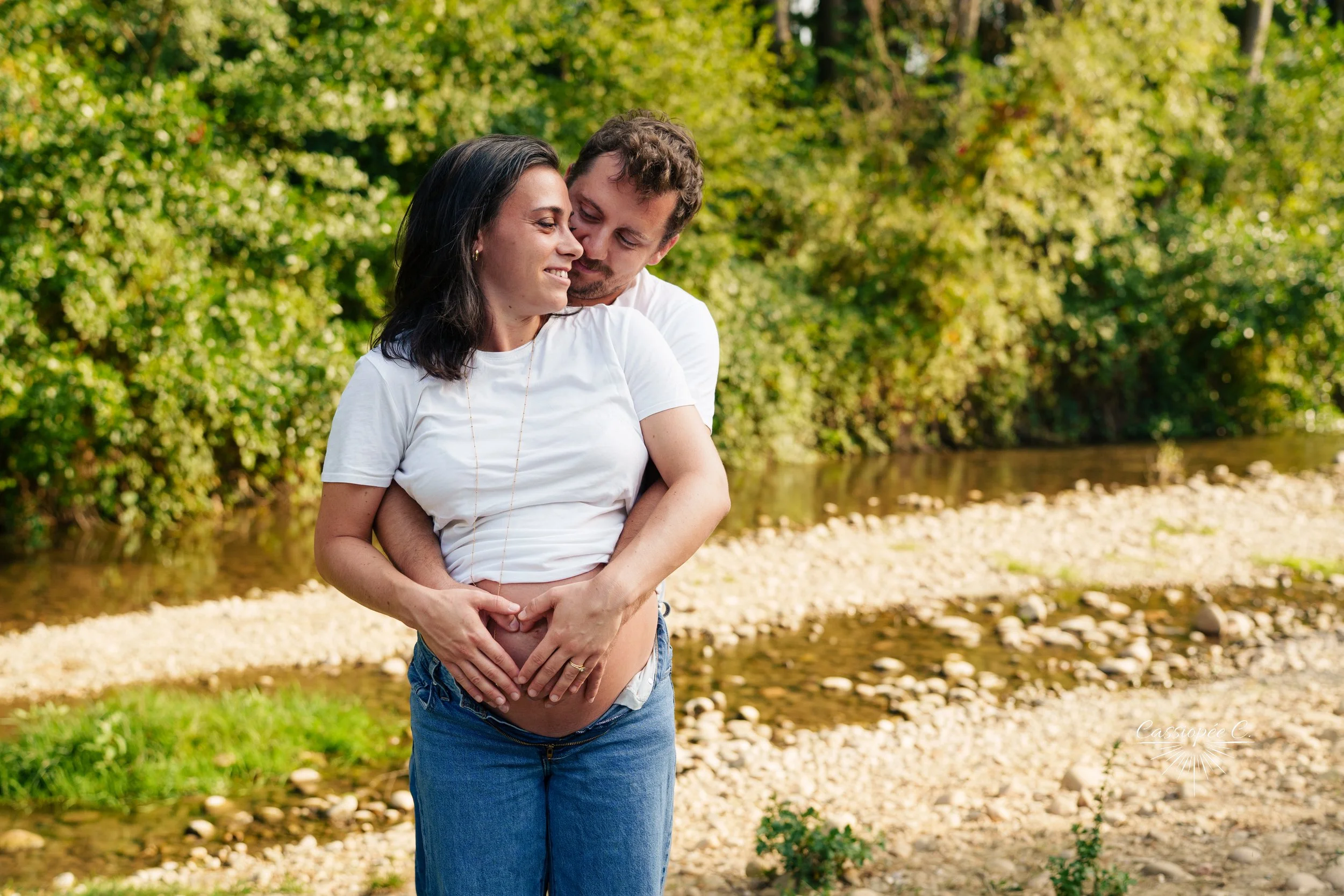 Un couple en balade près d'une rivière dans un parc, l'homme embrasse tendrement la femme est enceinte.