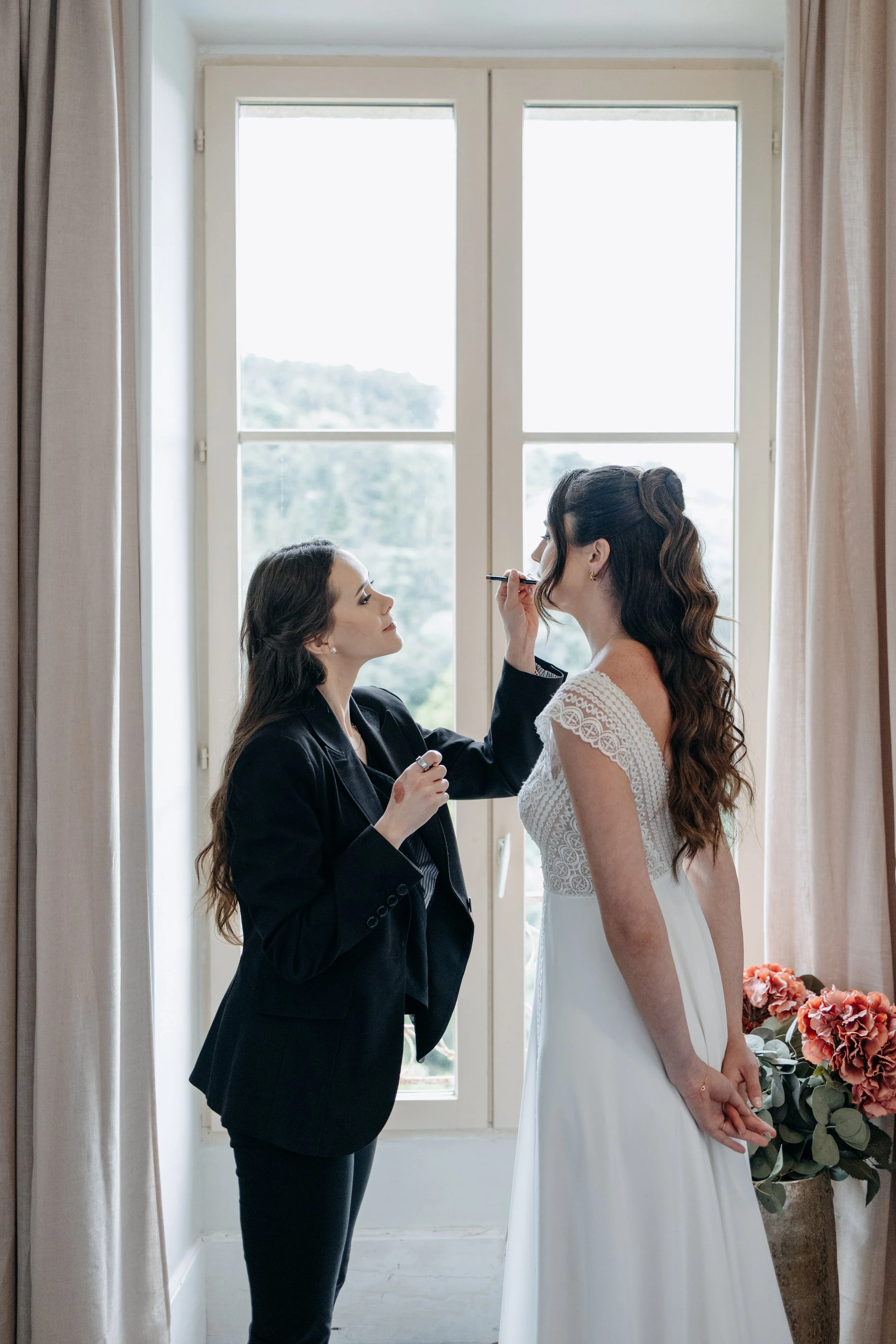 Une maquilleuse maquillant le visage d'une mariée, devant une grande fenêtre avec des rideaux beiges.