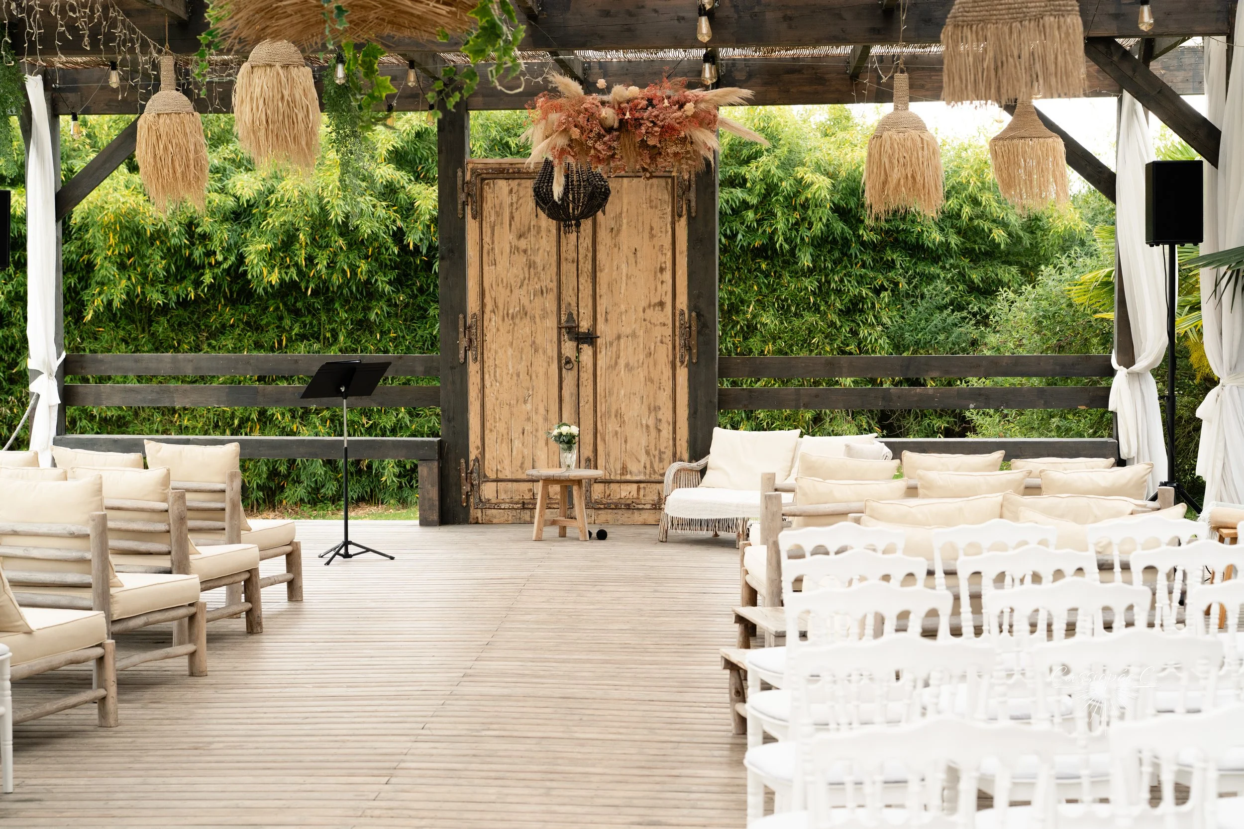 Espace de mariage en plein air avec sièges blancs et crème, décor bohème avec lanternes en paille suspendues, porte rustique en bois, et végétation verte en arrière-plan.