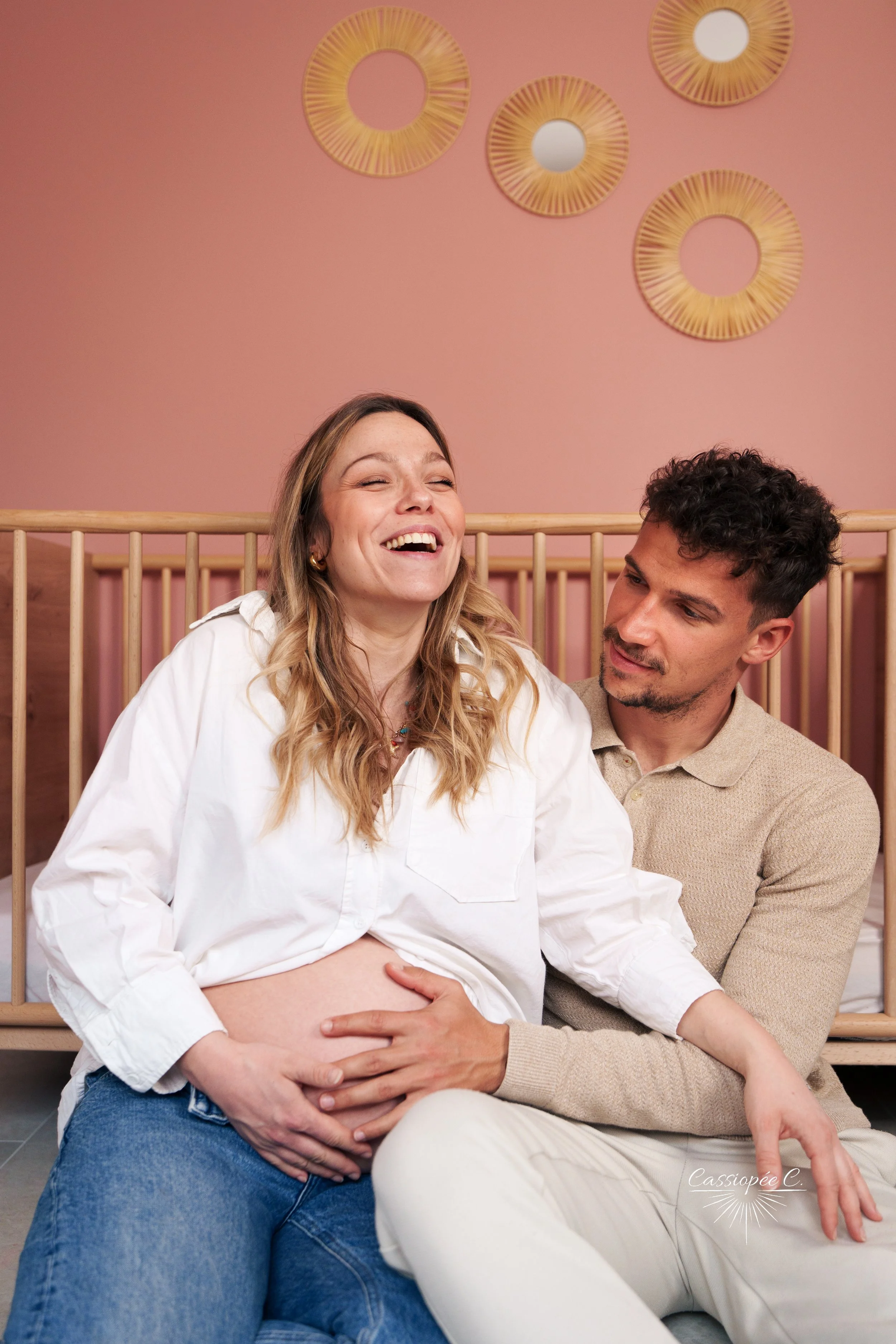 Une femme enceinte souriante assise sur les genoux d'un homme, devant un mur rose avec des décorations en miroir rondes.