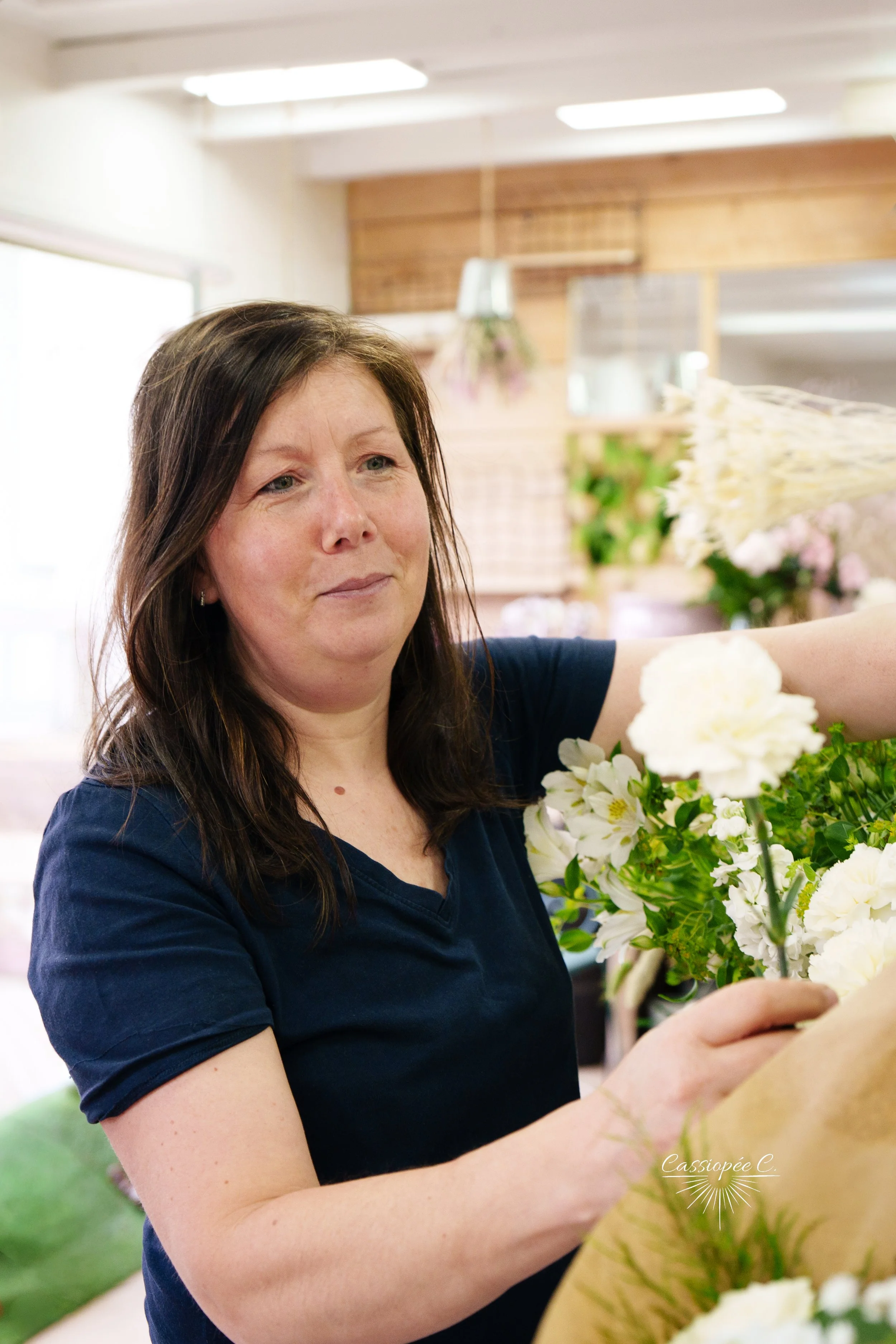 Une femme avec des cheveux bruns et un t-shirt bleu arrangeant un bouquet de fleurs blanches dans un magasin de fleurs.
