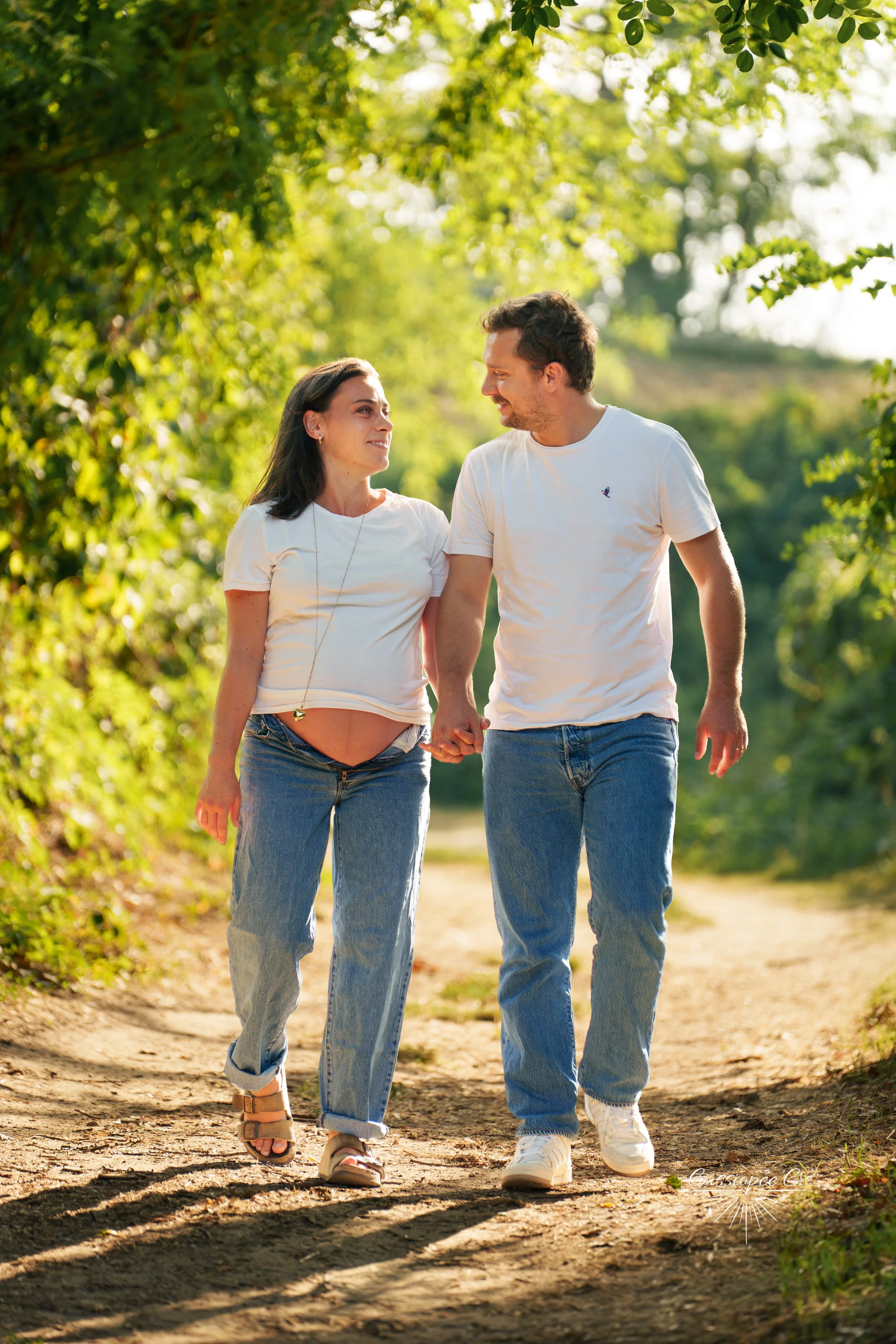 Un couple marche main dans la main sur un sentier en plein air, entouré de végétation luxuriante, lors d'une journée ensoleillée.