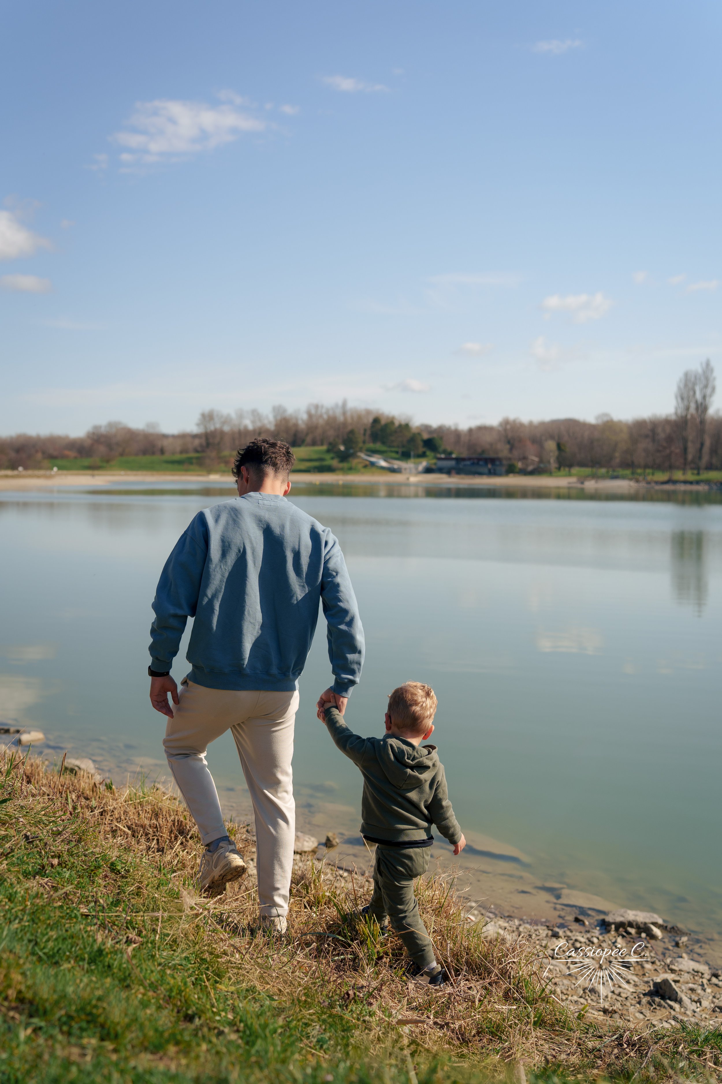 Un adulte et un enfant, bras liés, marche à côté d'un lac. Le ciel est bleu avec quelques nuages, et un paysage de collines et de bâtiments est visible en arrière-plan.