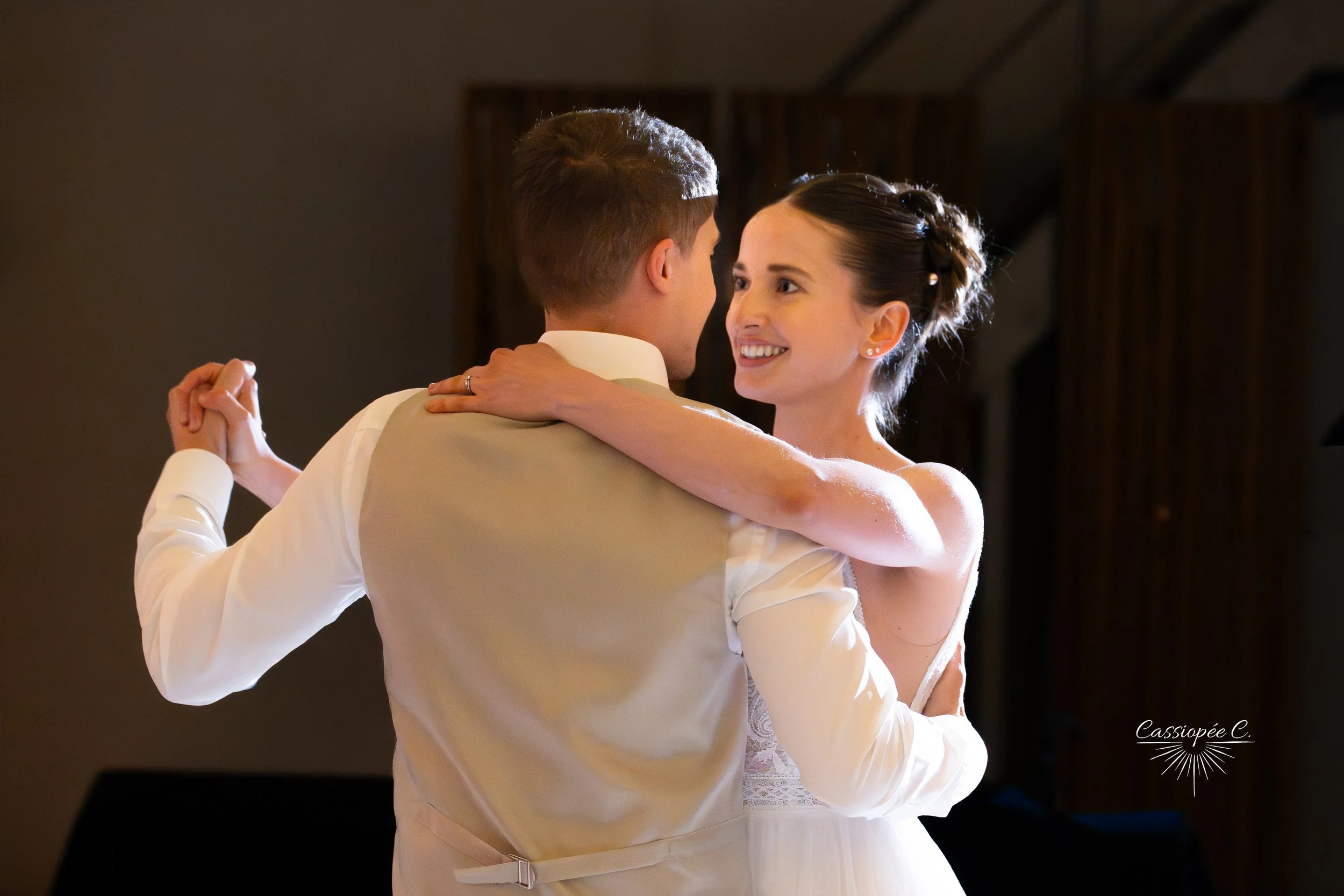 Un couple danse lors d'un mariage, la femme sourit alors qu'elle regarde son partenaire, ils portent des vêtements de mariage et se tiennent dans une pièce sombre avec un éclairage chaleureux.