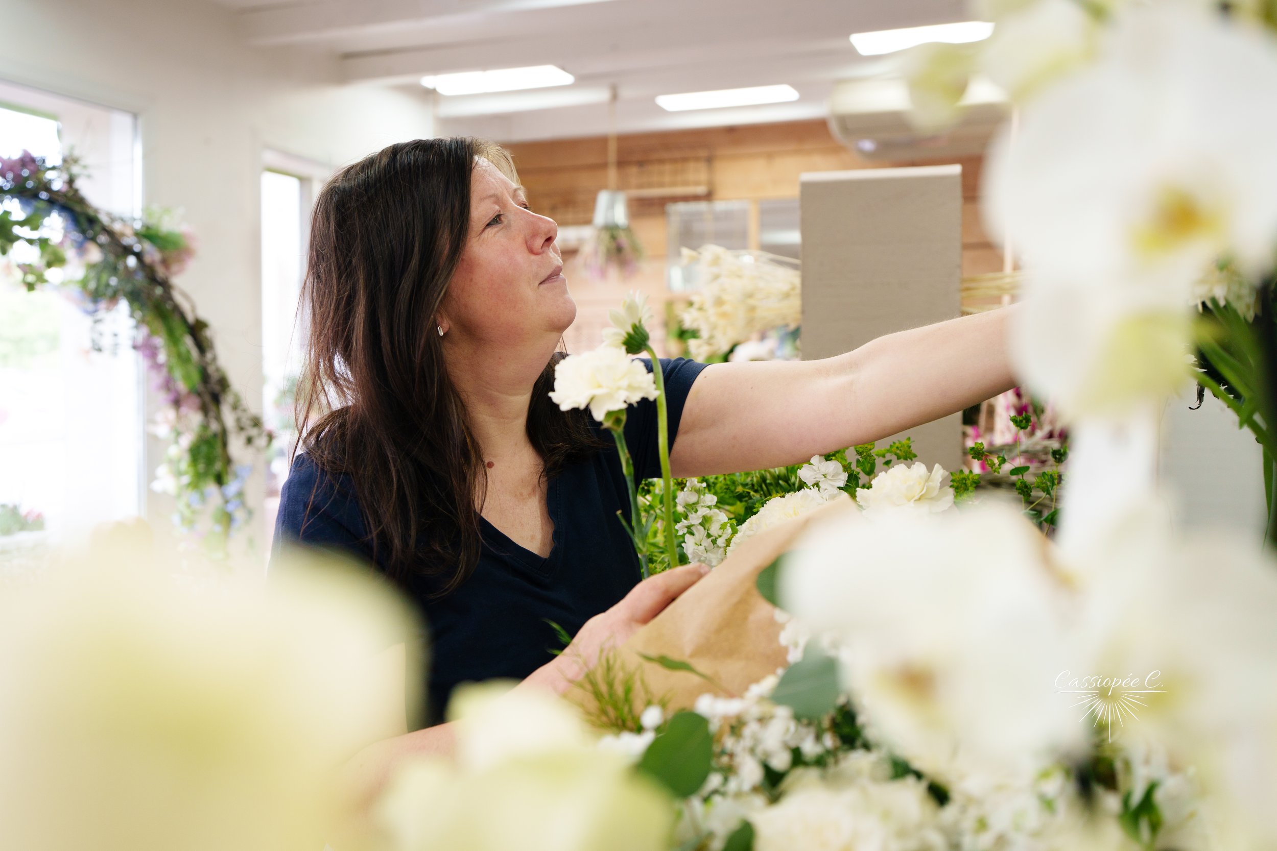 Une fleuriste avec les yeux fermés et un sourire léger, choisit des fleurs dans son magasin de fleurs, entourée de bouquets de fleurs blanches, dans un intérieur lumineux avec des fenêtres et des murs en bois.